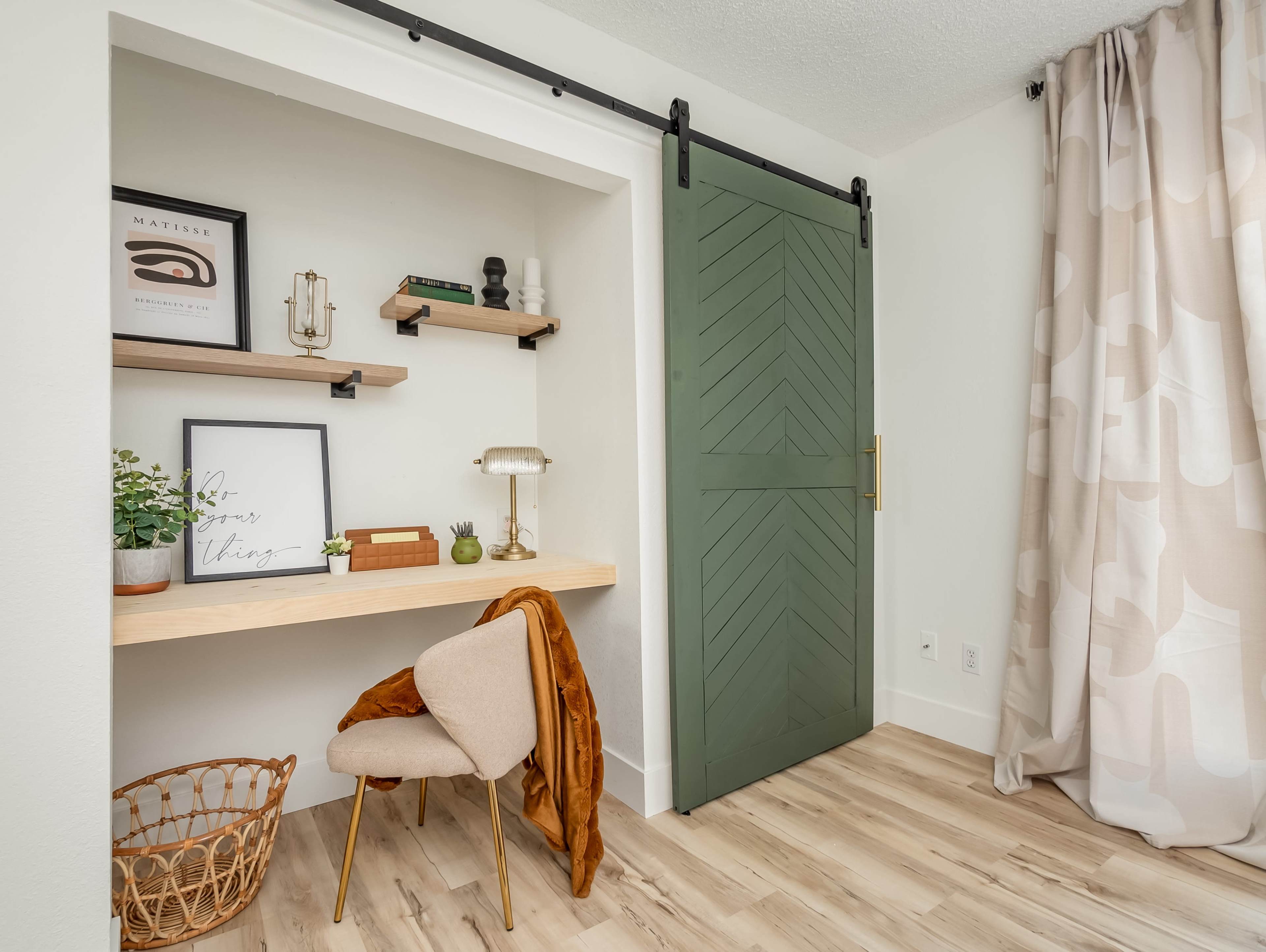 A modern interior corner featuring a sliding green barn door, a small wooden desk with decorative items, and a chair beside a woven basket.