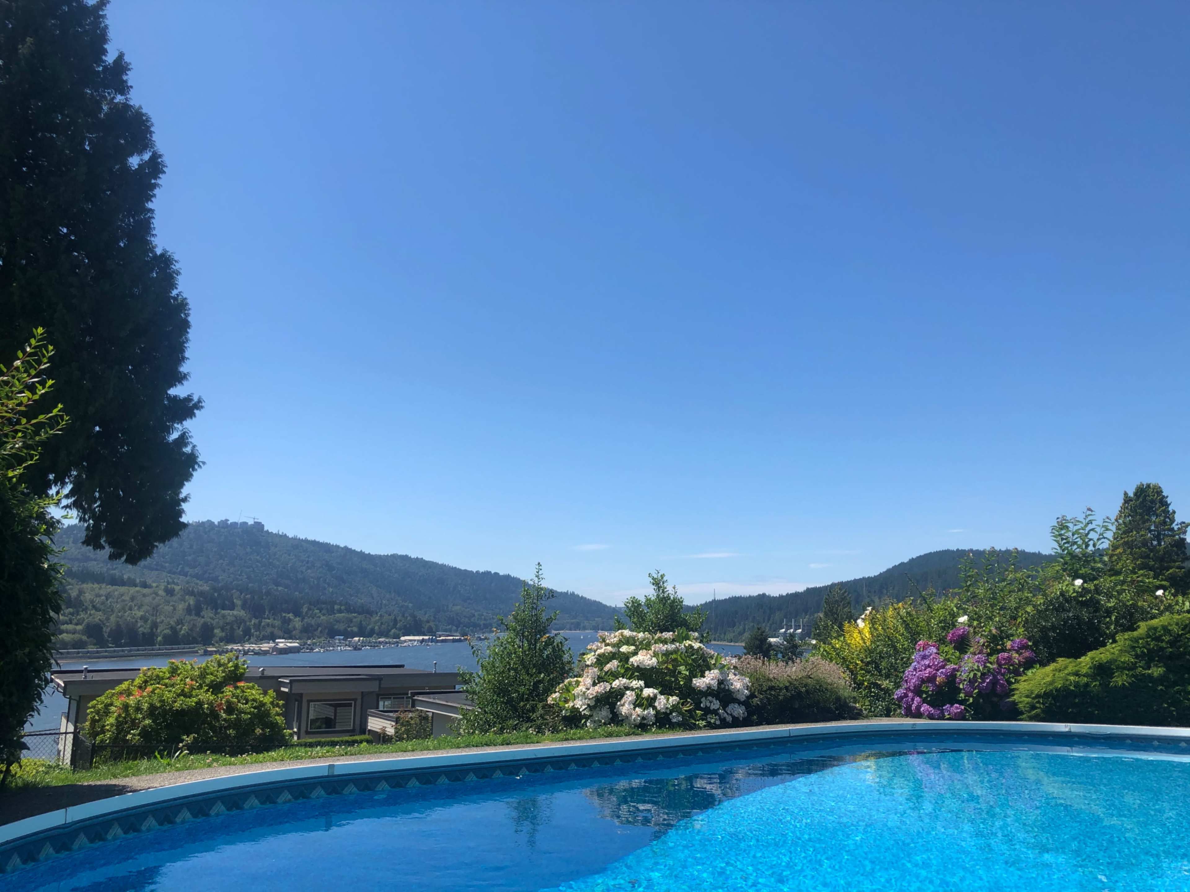 A swimming pool is surrounded by greenery and flowers, overlooking a lake with hills in the background under a clear blue sky.