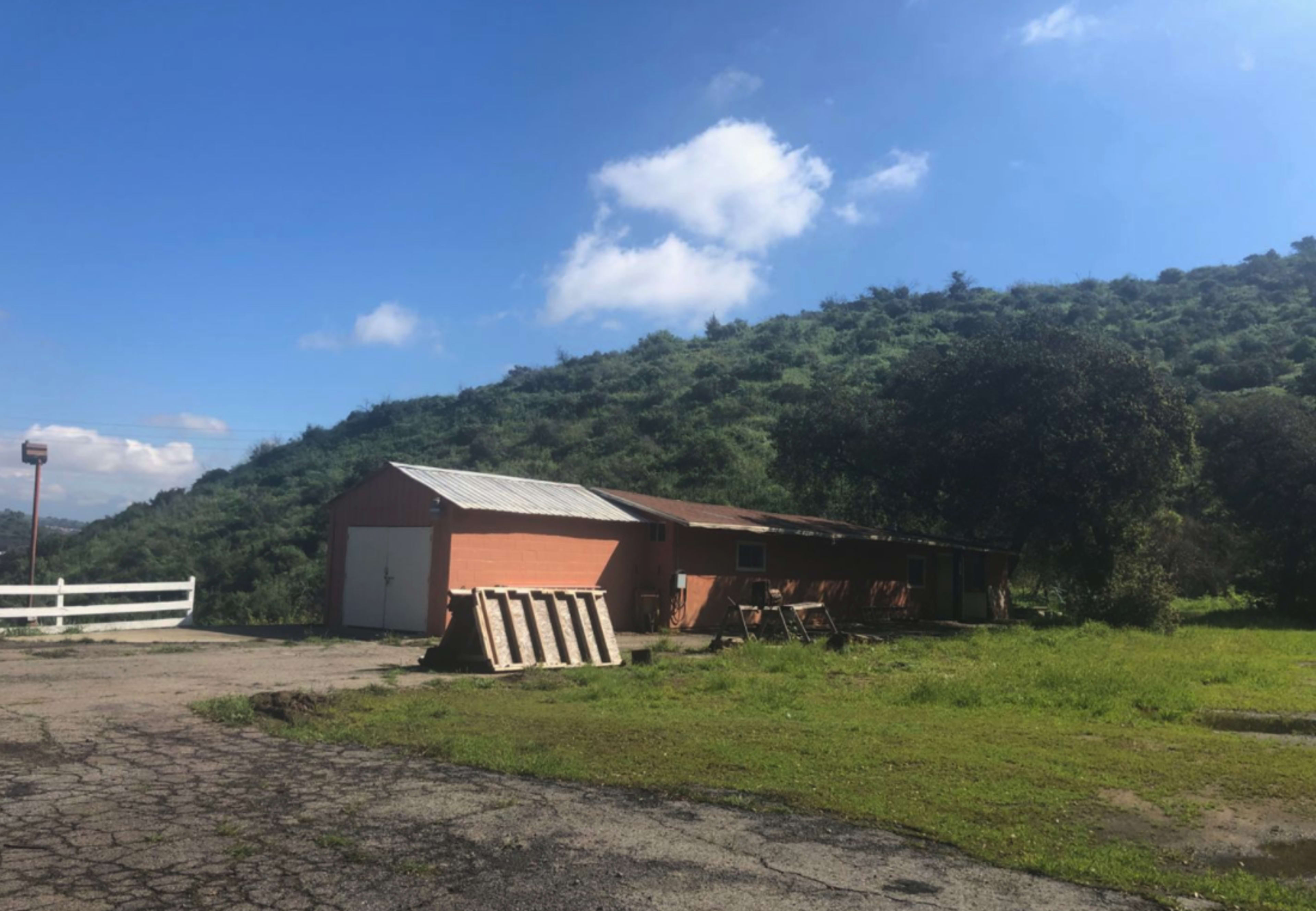 A single-story orange building with a metal roof is situated near a grassy area and a low hill under a clear blue sky.