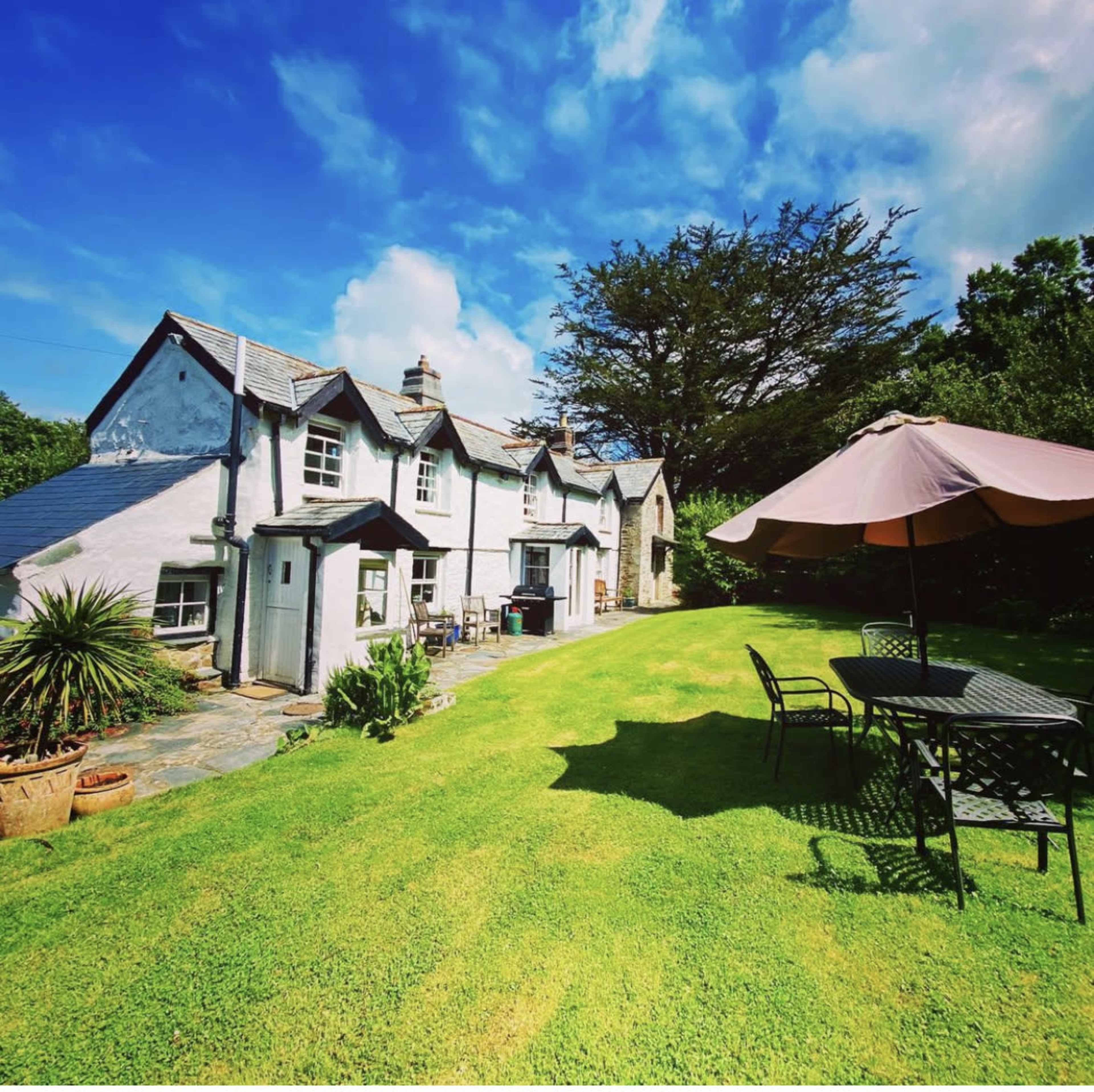 A cottage with a slate roof sits in a green garden, accompanied by a patio table and umbrella under a clear blue sky.