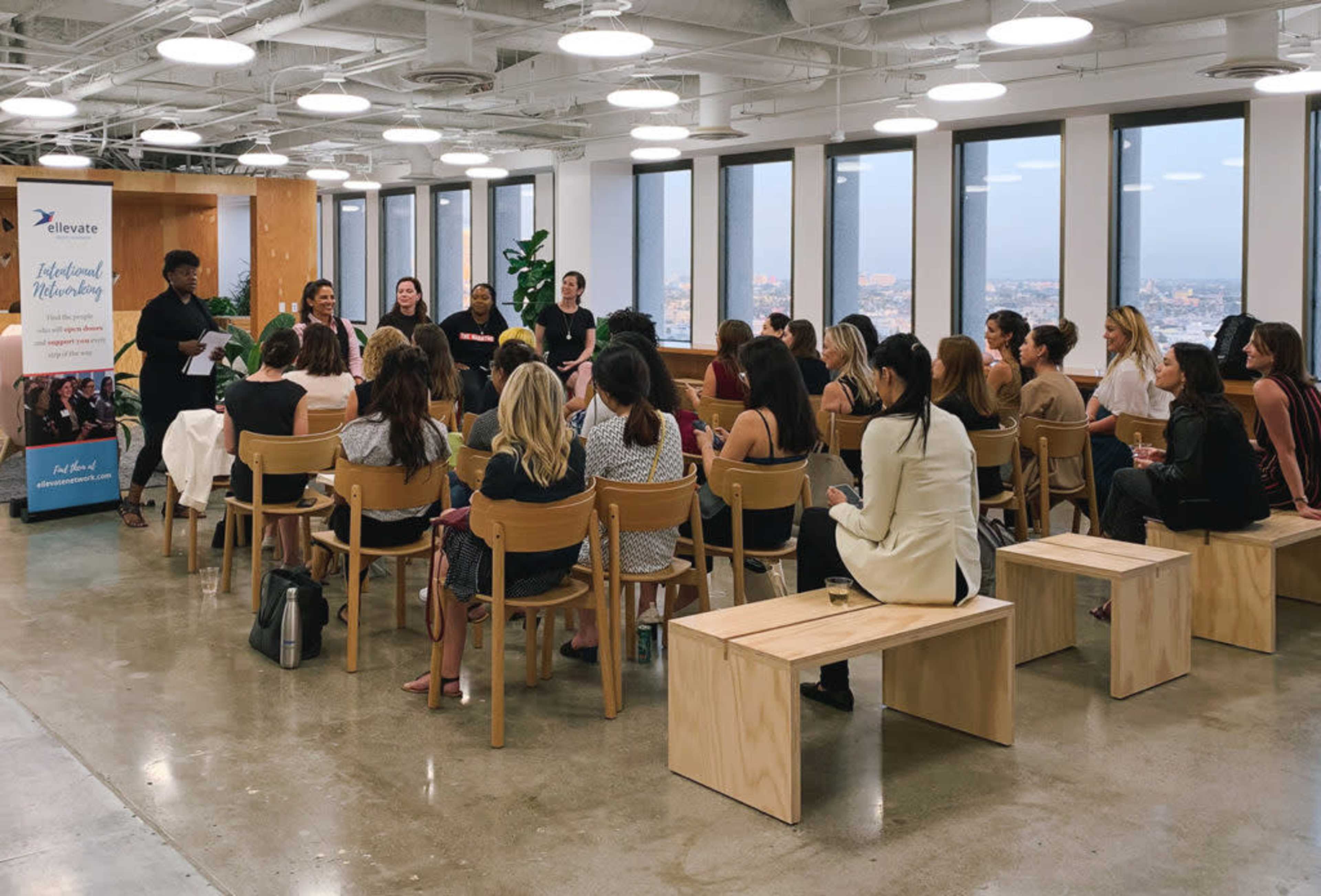 A group of women is seated in a modern office space, attending a presentation or discussion by several speakers in front of large windows showcasing a city view.