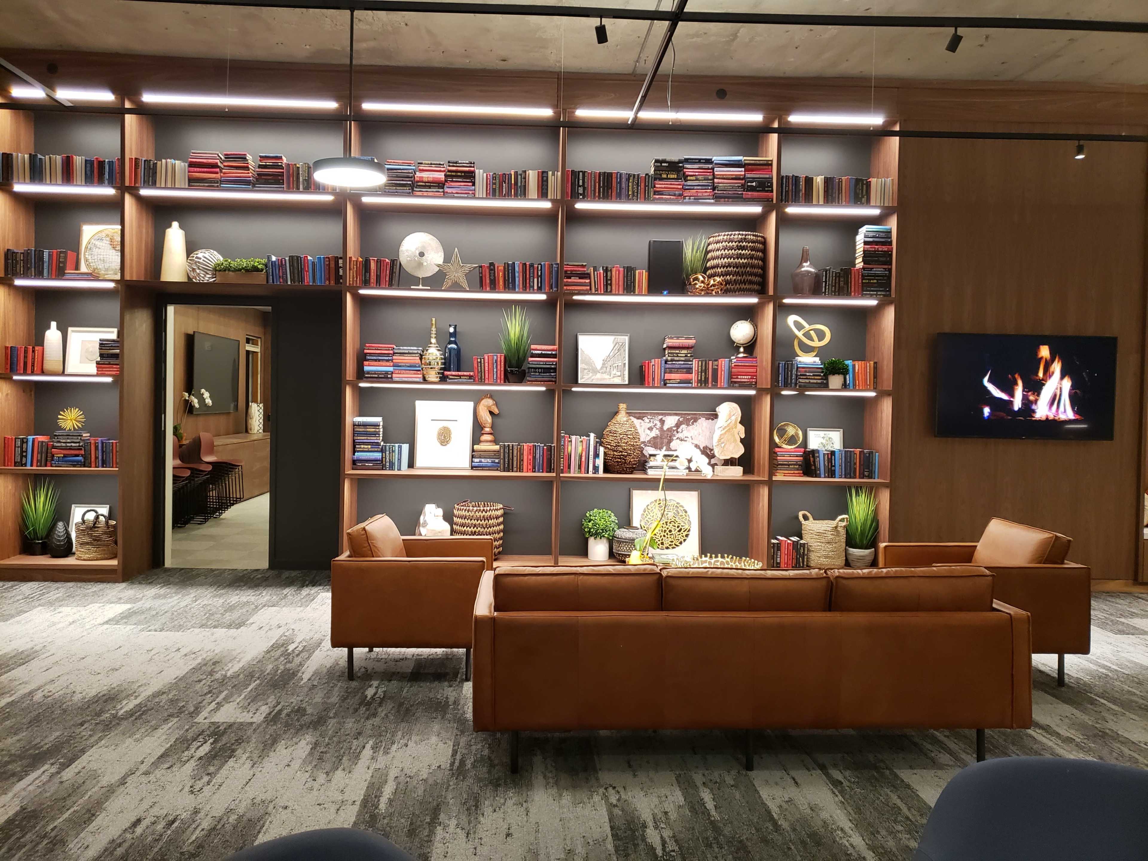 A modern lounge area featuring a brown leather sofa facing a large bookshelf filled with books and decorative items, with a television displaying a fireplace on one side.