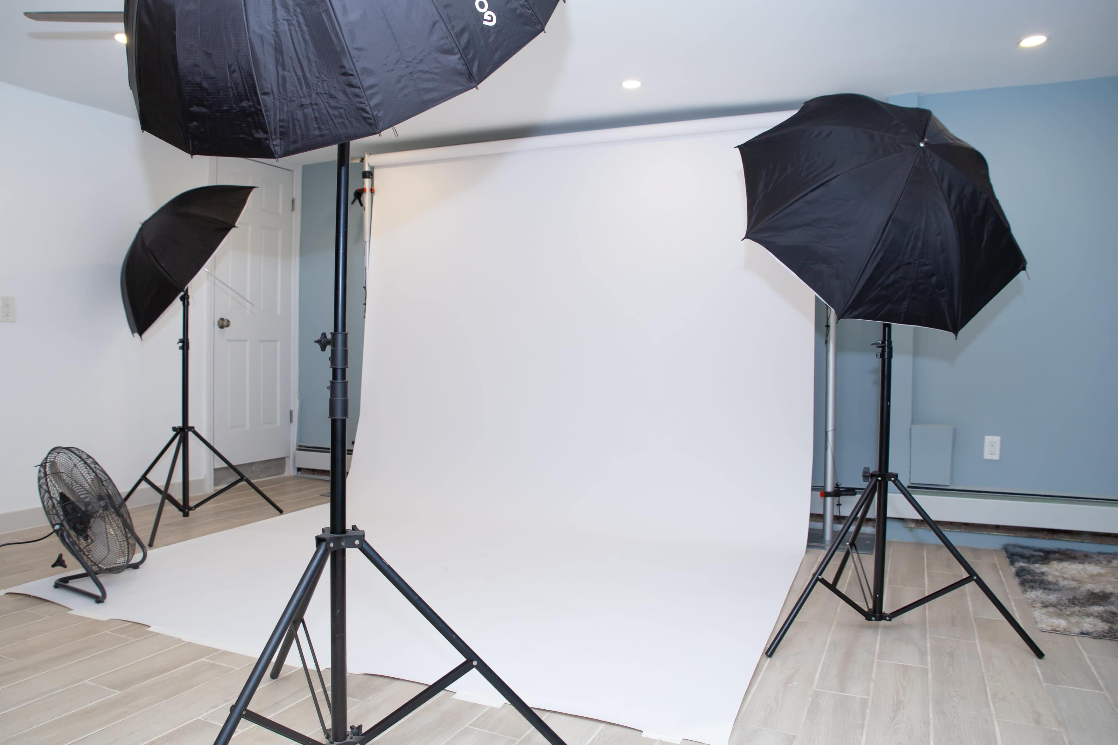 The image shows a photography studio setup with three light umbrellas and a backdrop of white fabric.