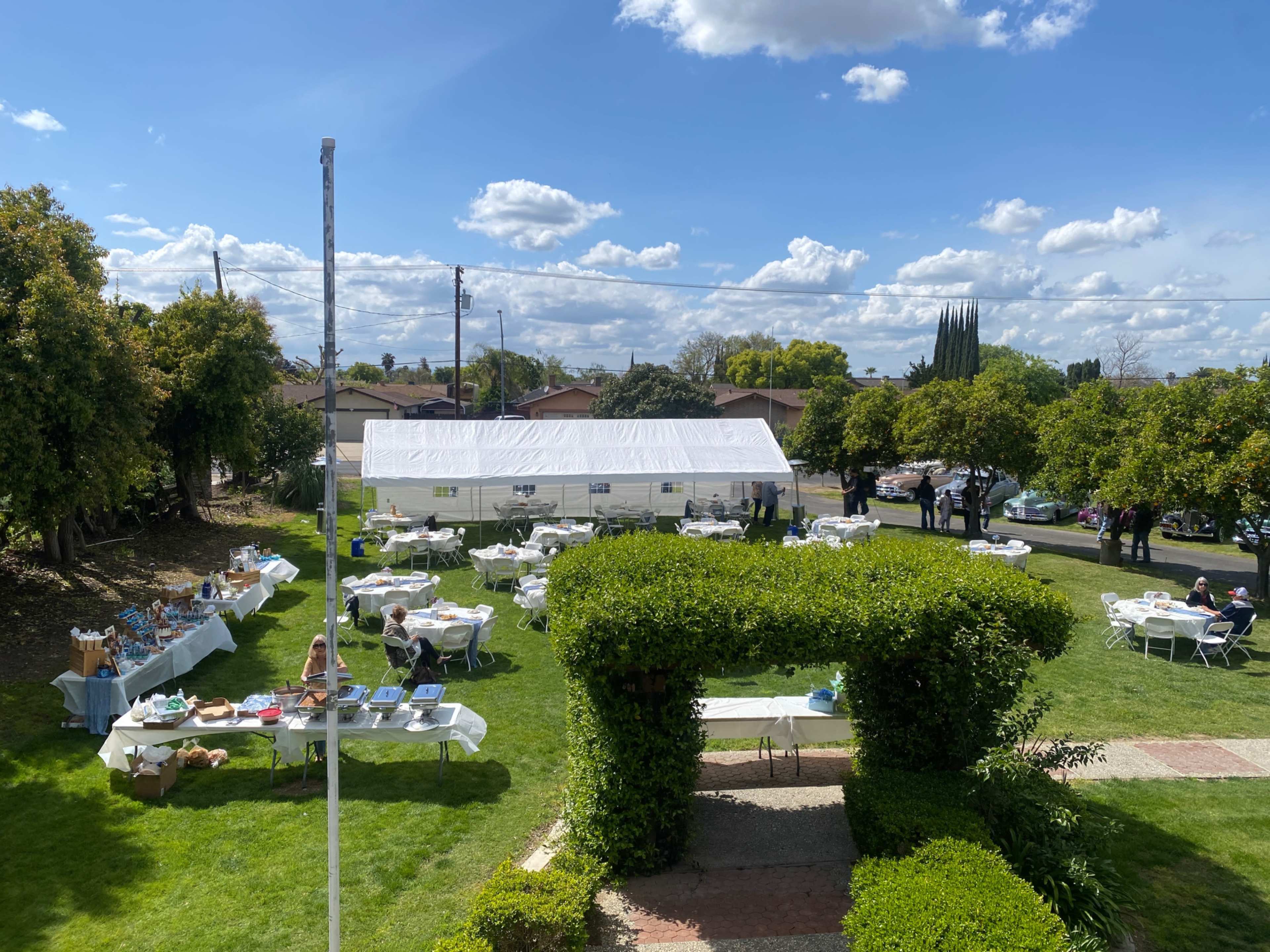 The image shows a large outdoor event with numerous tables set up under a white tent on a grassy area, surrounded by trees and blue skies with clouds.