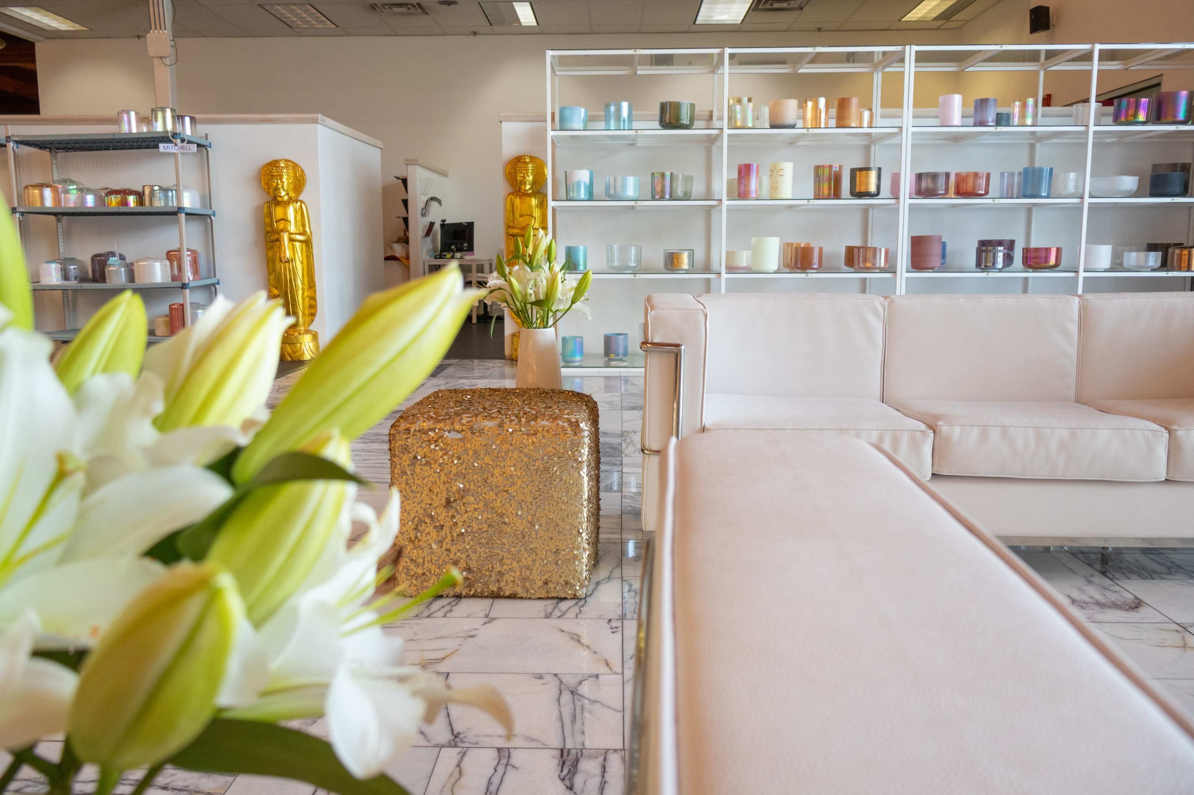 The image shows a modern retail space featuring gold statues, colorful candles on shelves, and white furniture complemented by a floral arrangement.