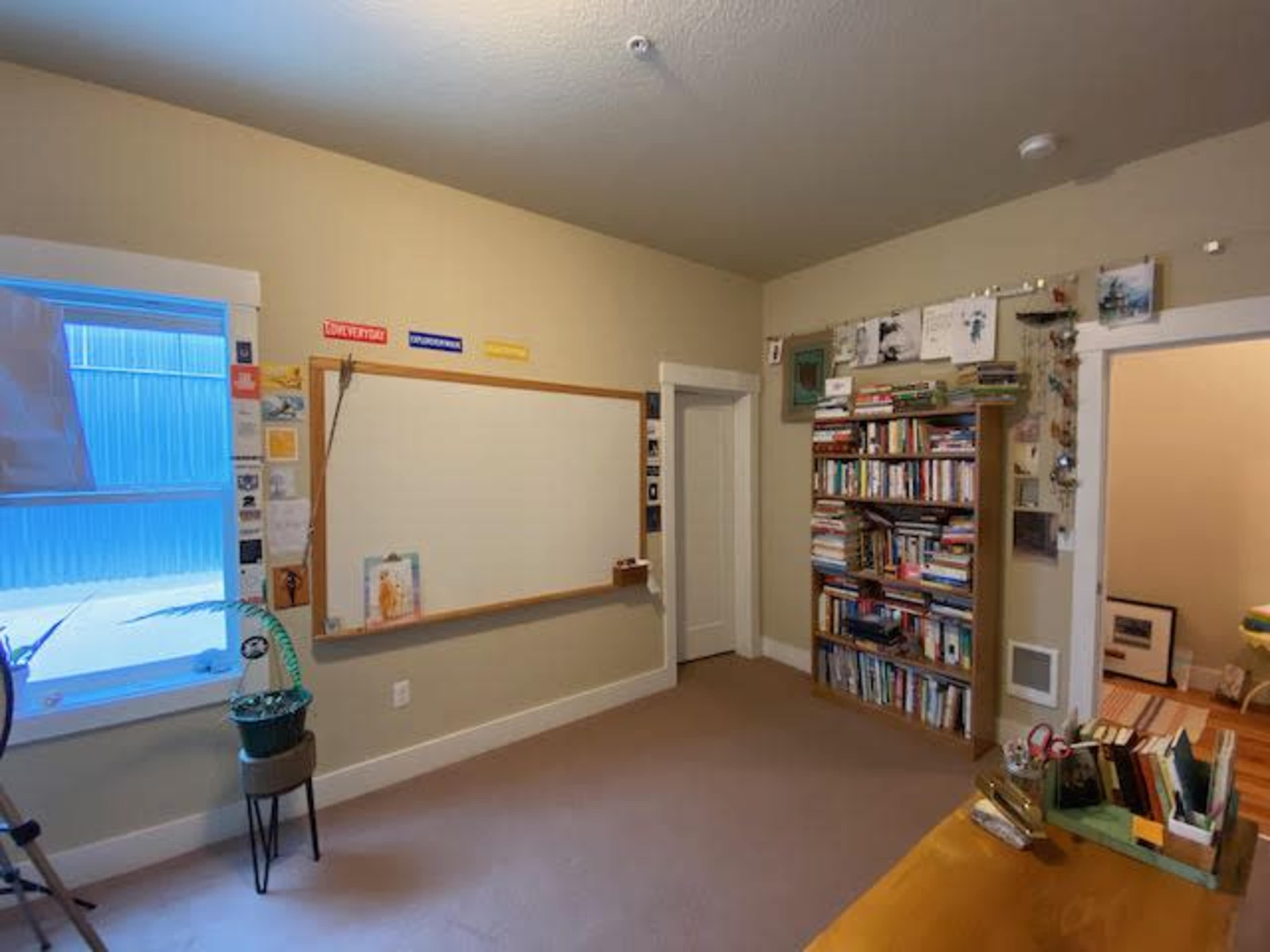 A room with a bookshelf filled with books, a blank whiteboard on the wall, and a window with blue curtains.