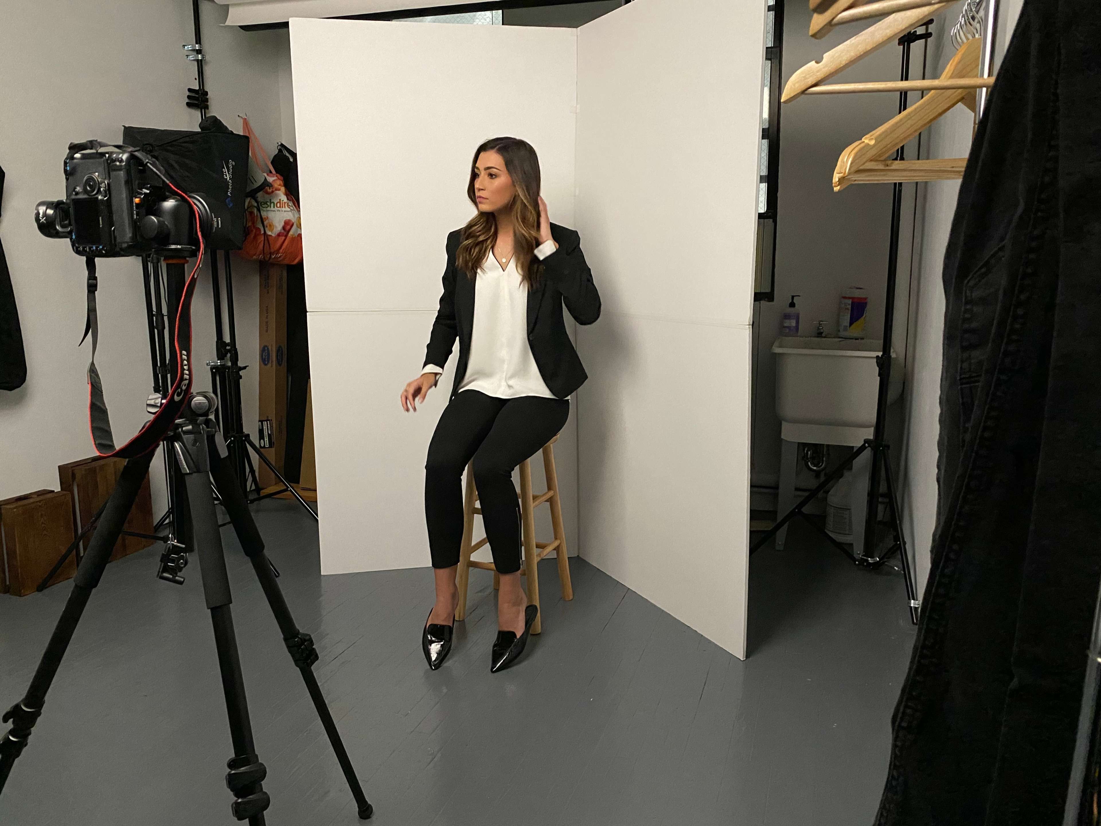 A woman in a black blazer and high heels is sitting on a stool in a photography studio, posing for the camera.