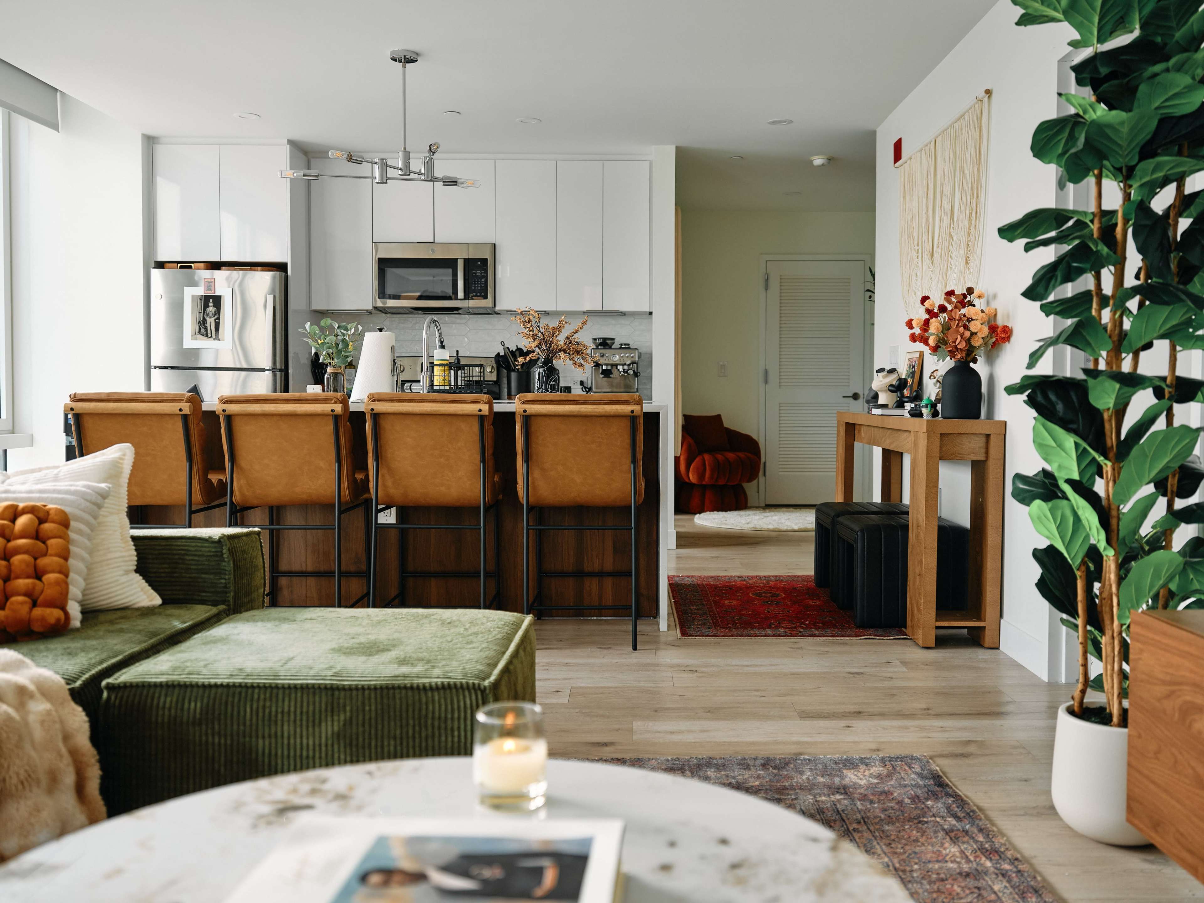 The image shows a modern open-concept living space featuring a kitchen with bar stools, a green sofa, and decorative plants.