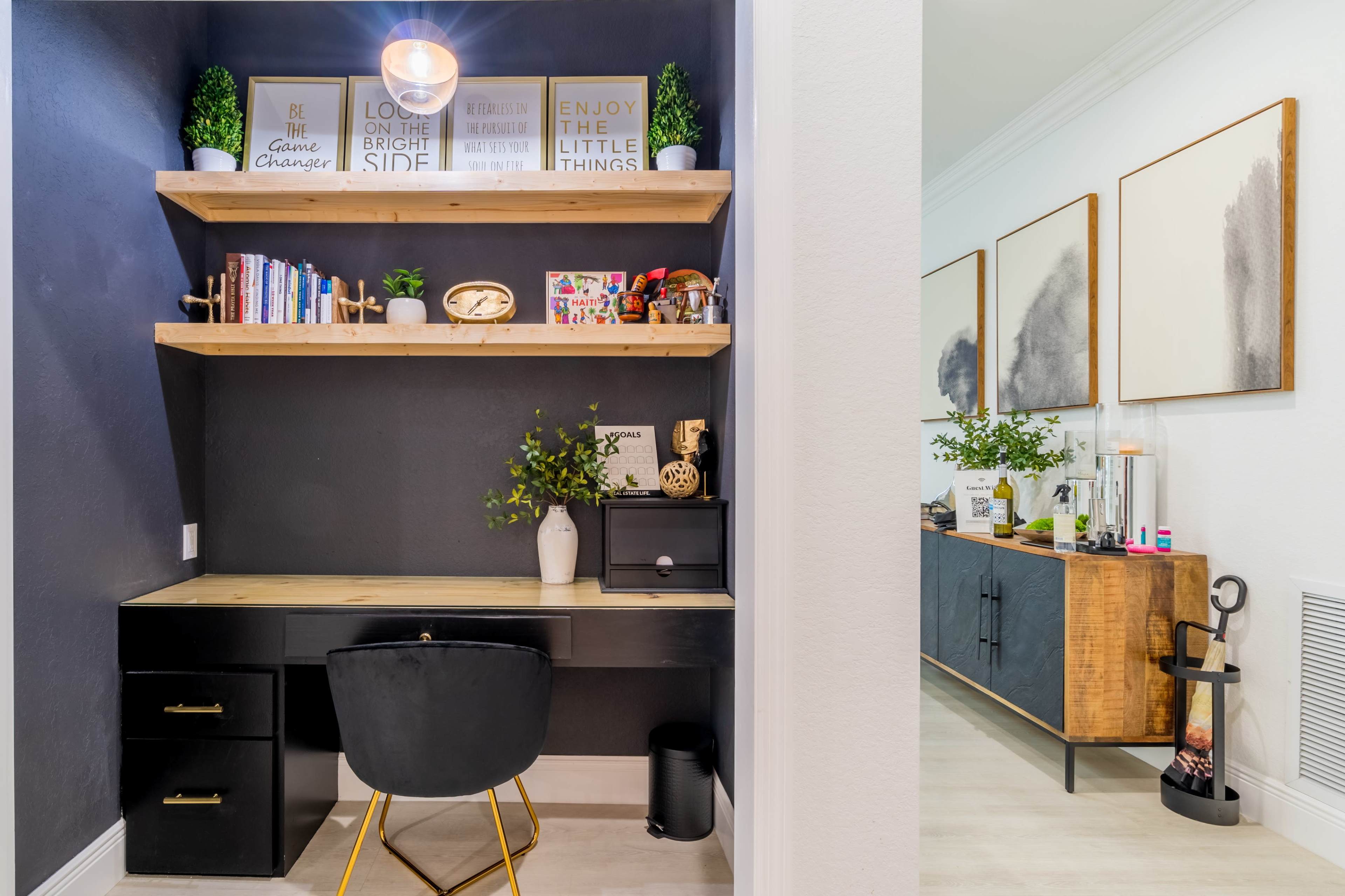 A modern home office setup with a wooden desk, shelves filled with books and decorative items, and a stylish chair beside a hallway featuring a sideboard and wall art.