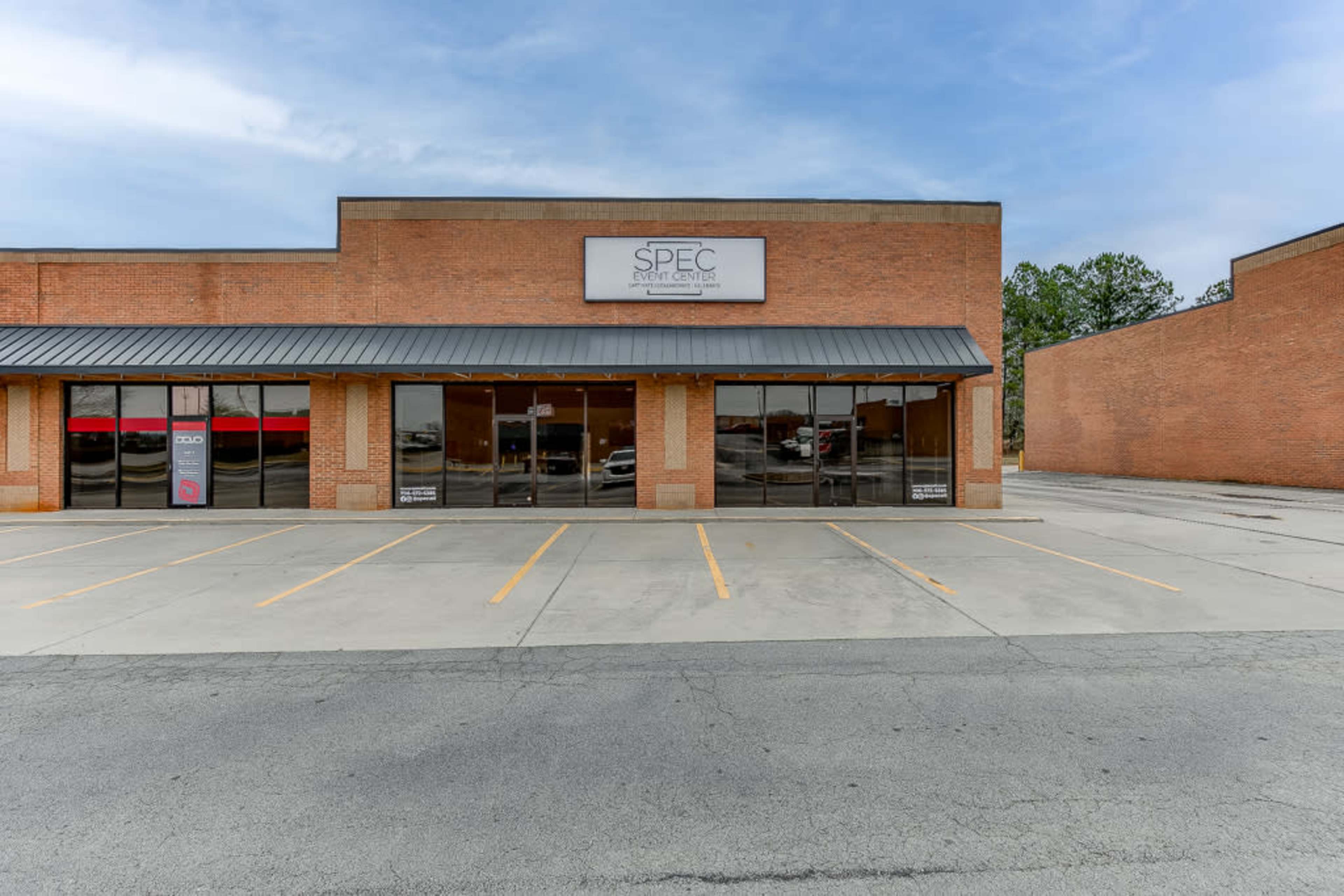 A brick commercial building with a sign for "SPEC" above large glass windows, situated in an empty parking lot.