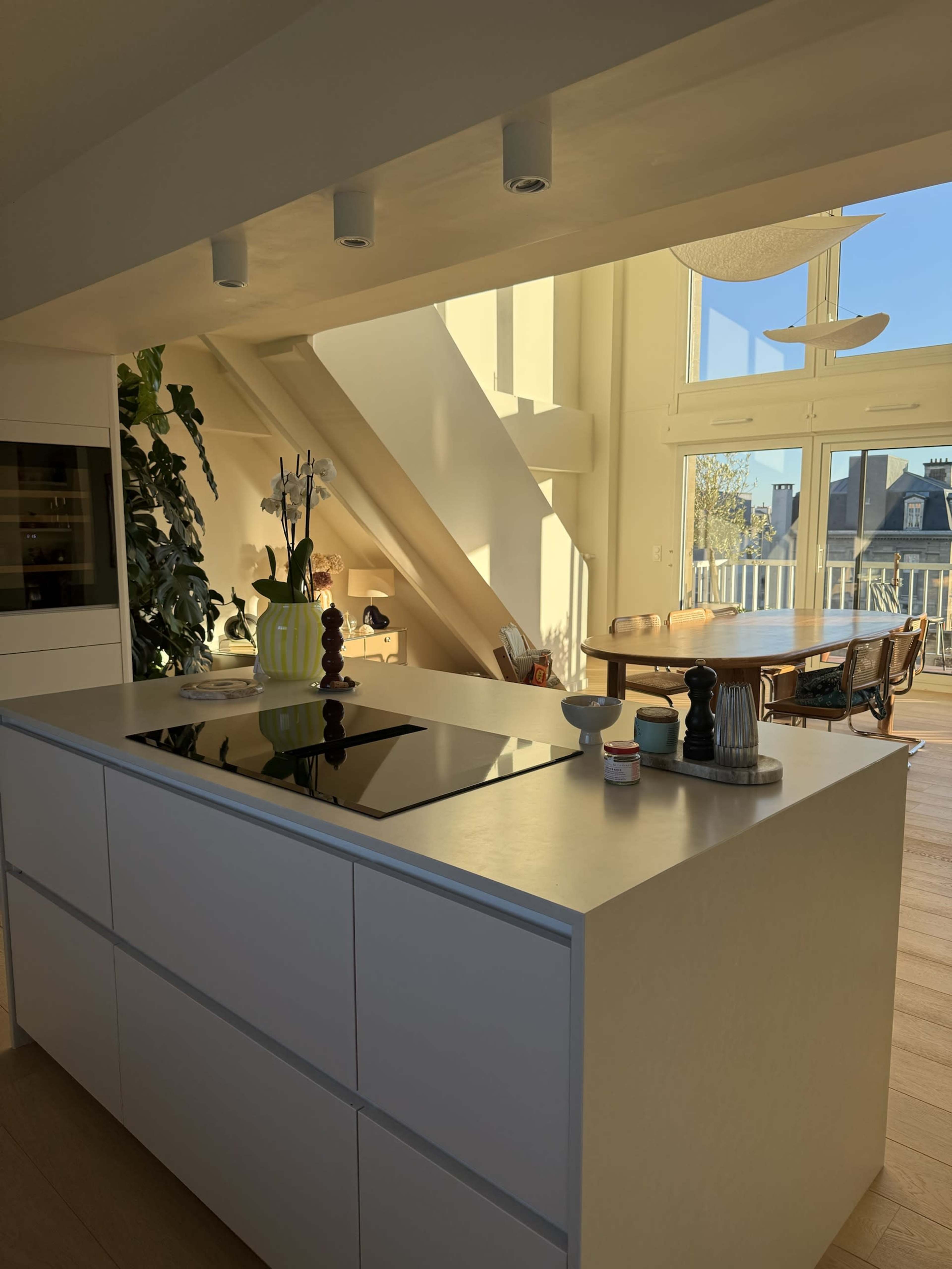 The photo shows a modern kitchen with a sleek island, featuring a stovetop and surrounded by natural light, and a dining area with wooden furniture in the background.