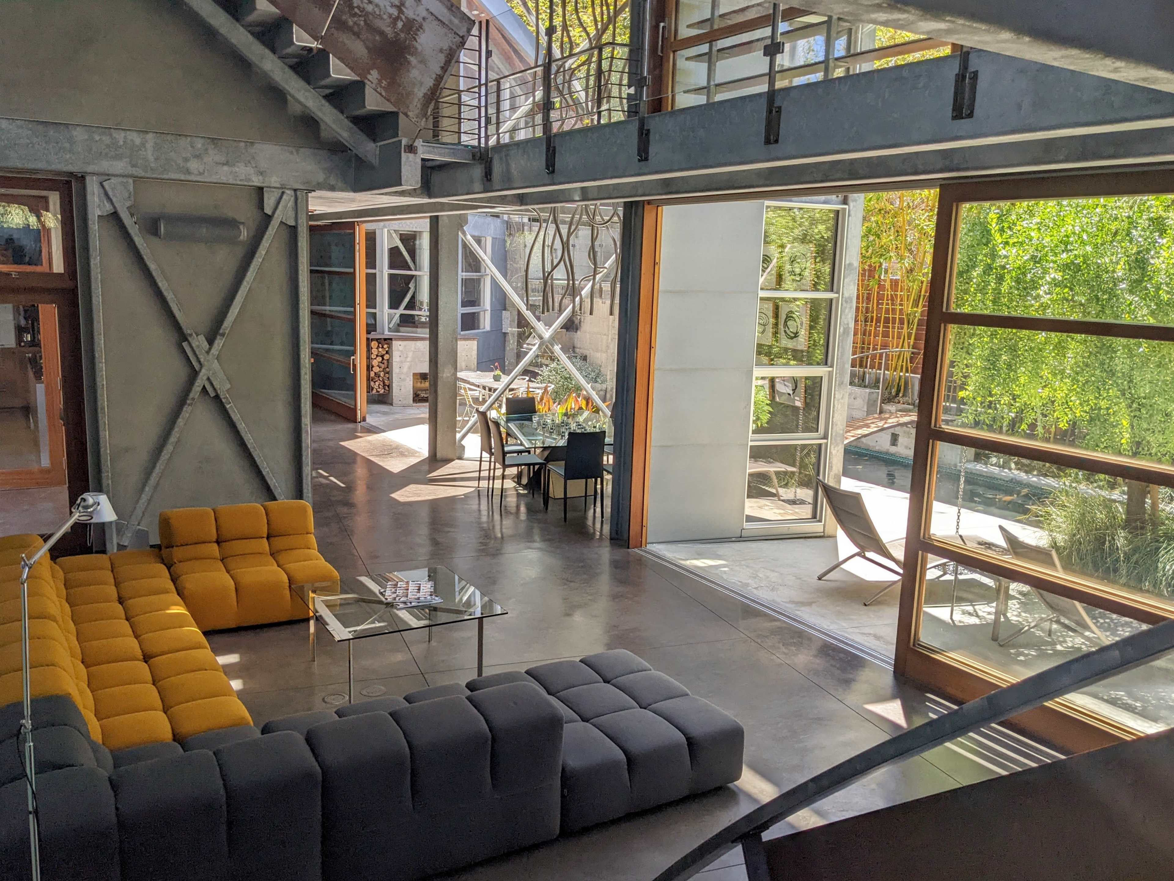 The image shows a modern living area with a gray and yellow sectional sofa, large glass walls, and a view of an outdoor space featuring a pool and seating.