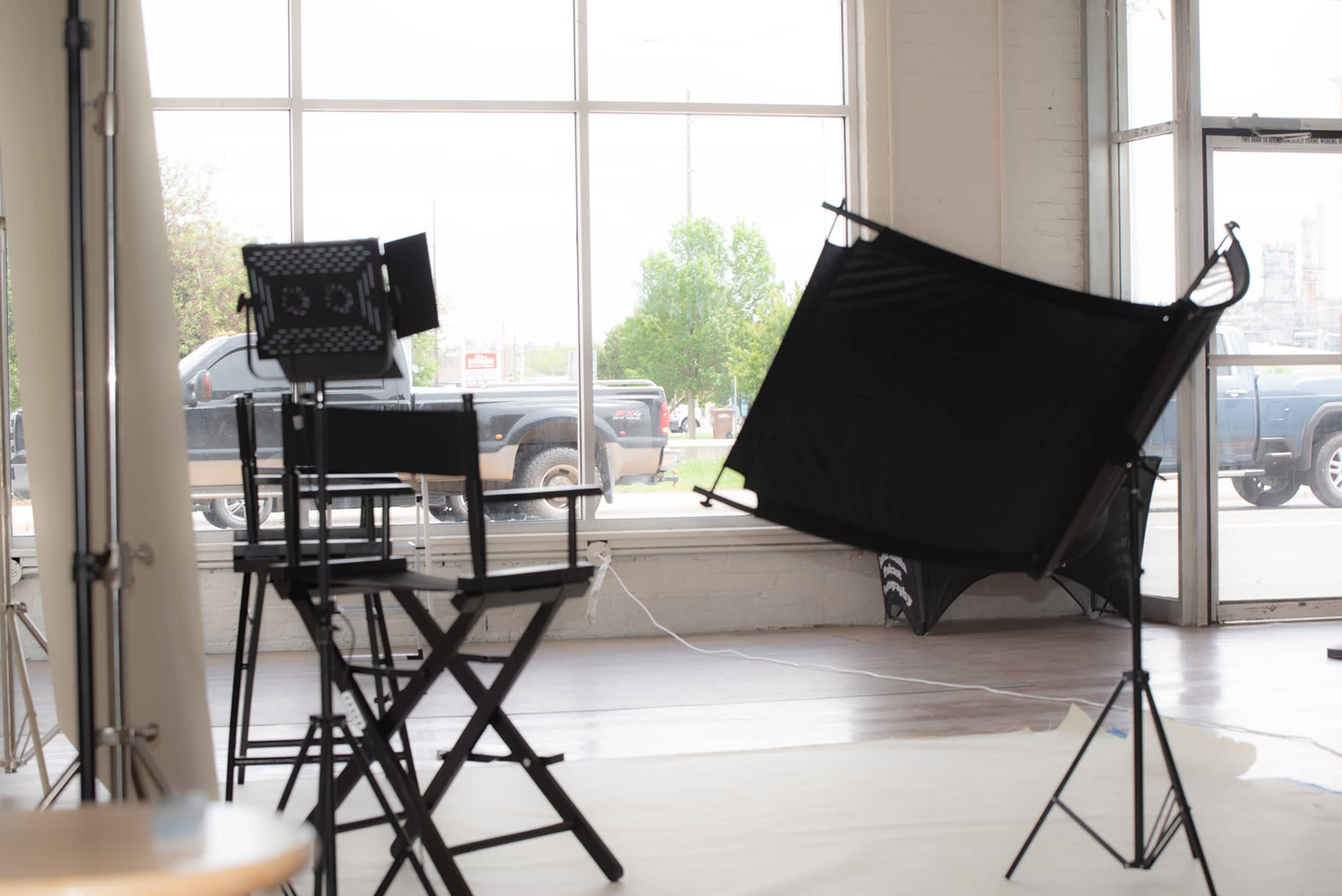 The image shows a photography studio with a large window, two director's chairs, a light setup, and a black backdrop positioned in front of the window.