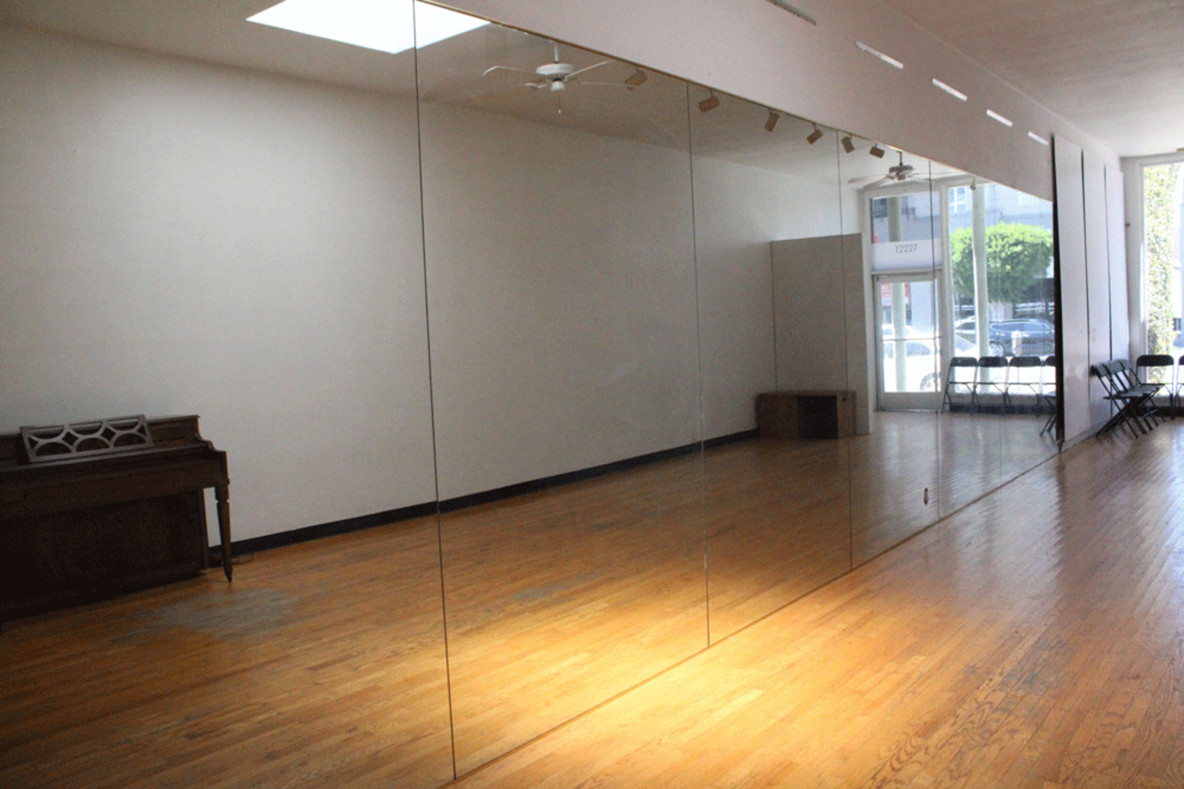 The image shows a spacious, empty room with a large mirror on one wall and wooden flooring, along with a piano and chairs set against the opposite wall.