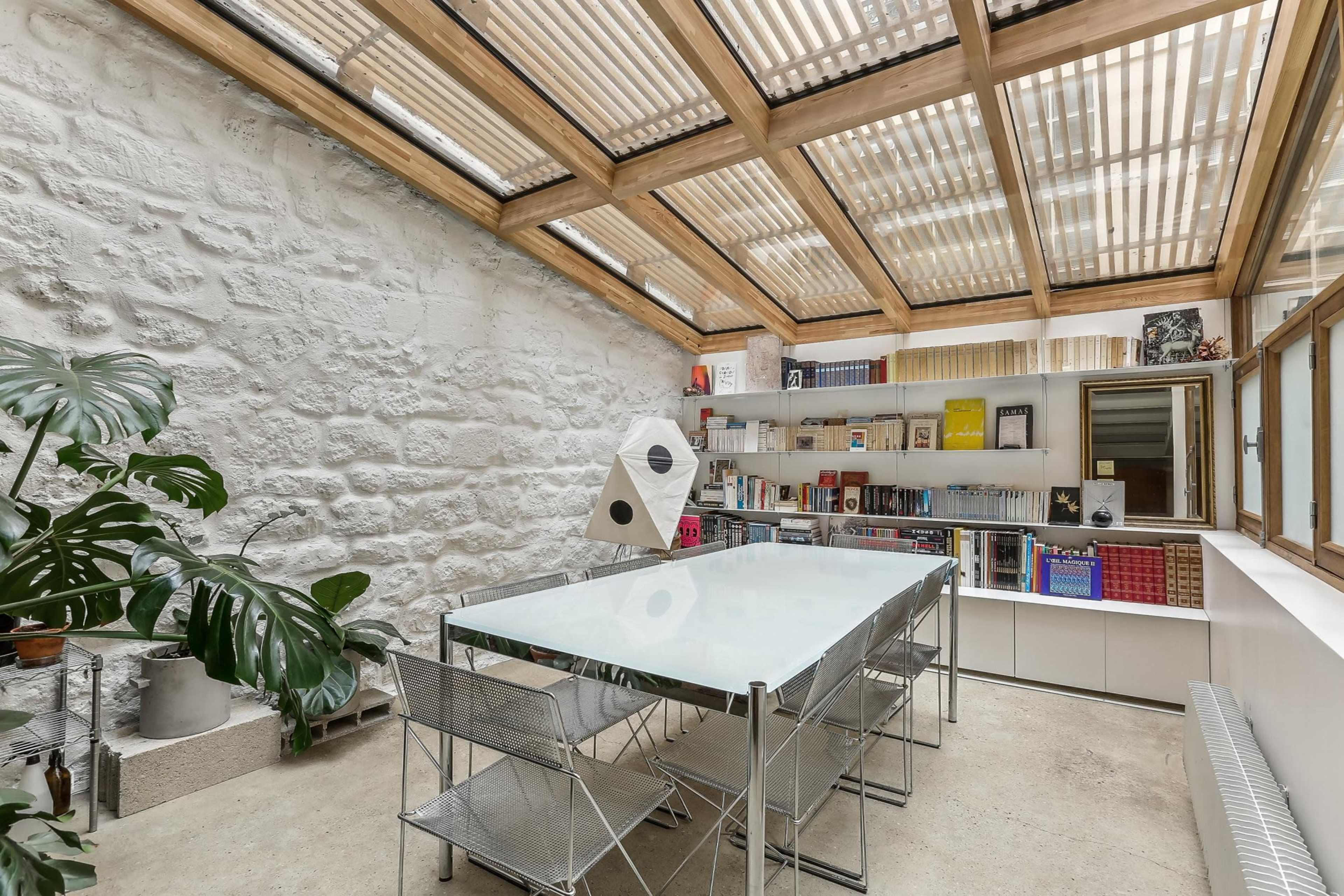The image depicts a modern study or office space with a glass table surrounded by metal chairs, bookshelves filled with various items, and a large skylight allowing natural light to enter.