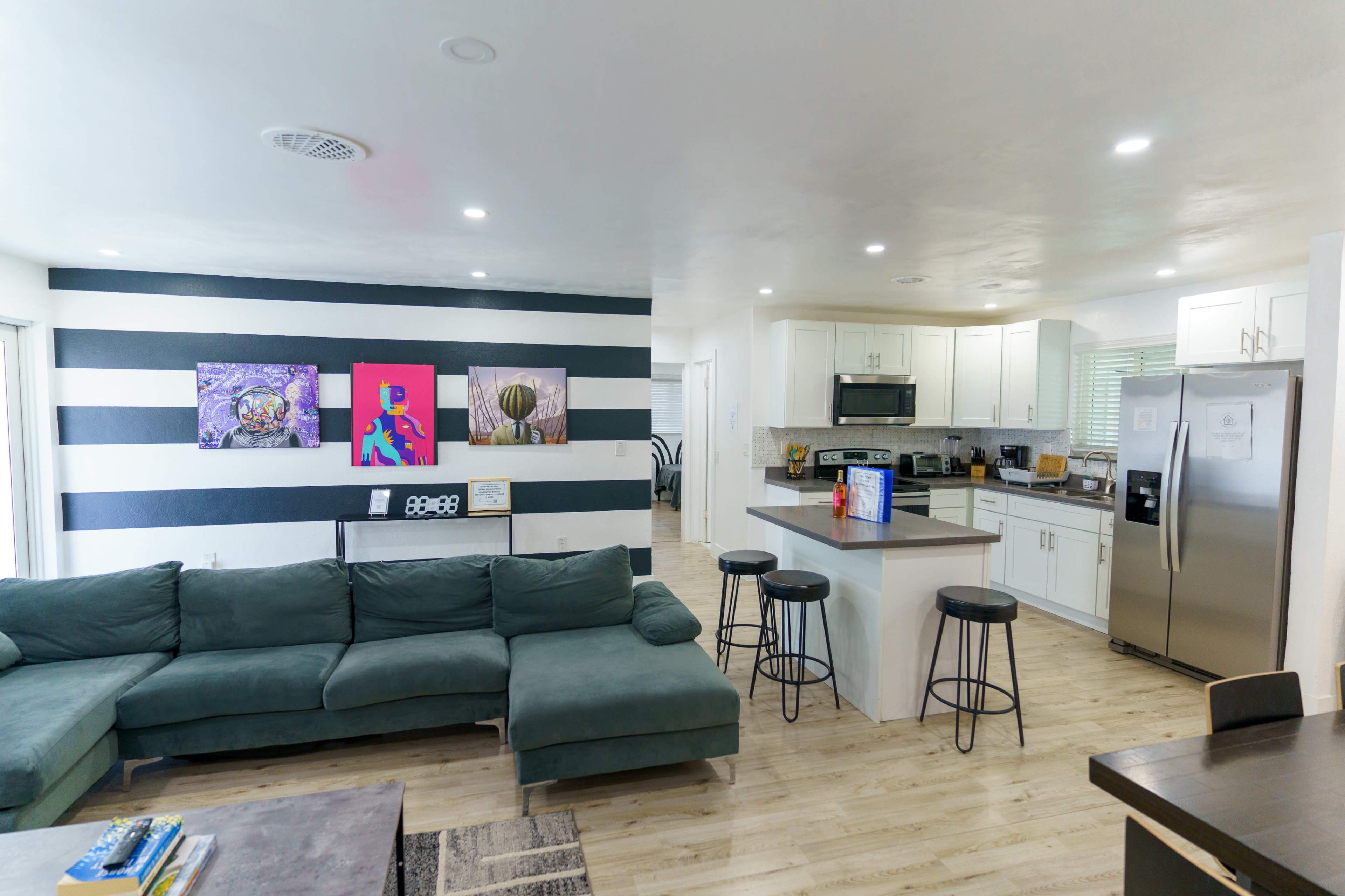 A modern open-concept living area features a green sectional sofa, black bar stools, and a kitchen with white cabinets and stainless steel appliances, complemented by colorful wall art.