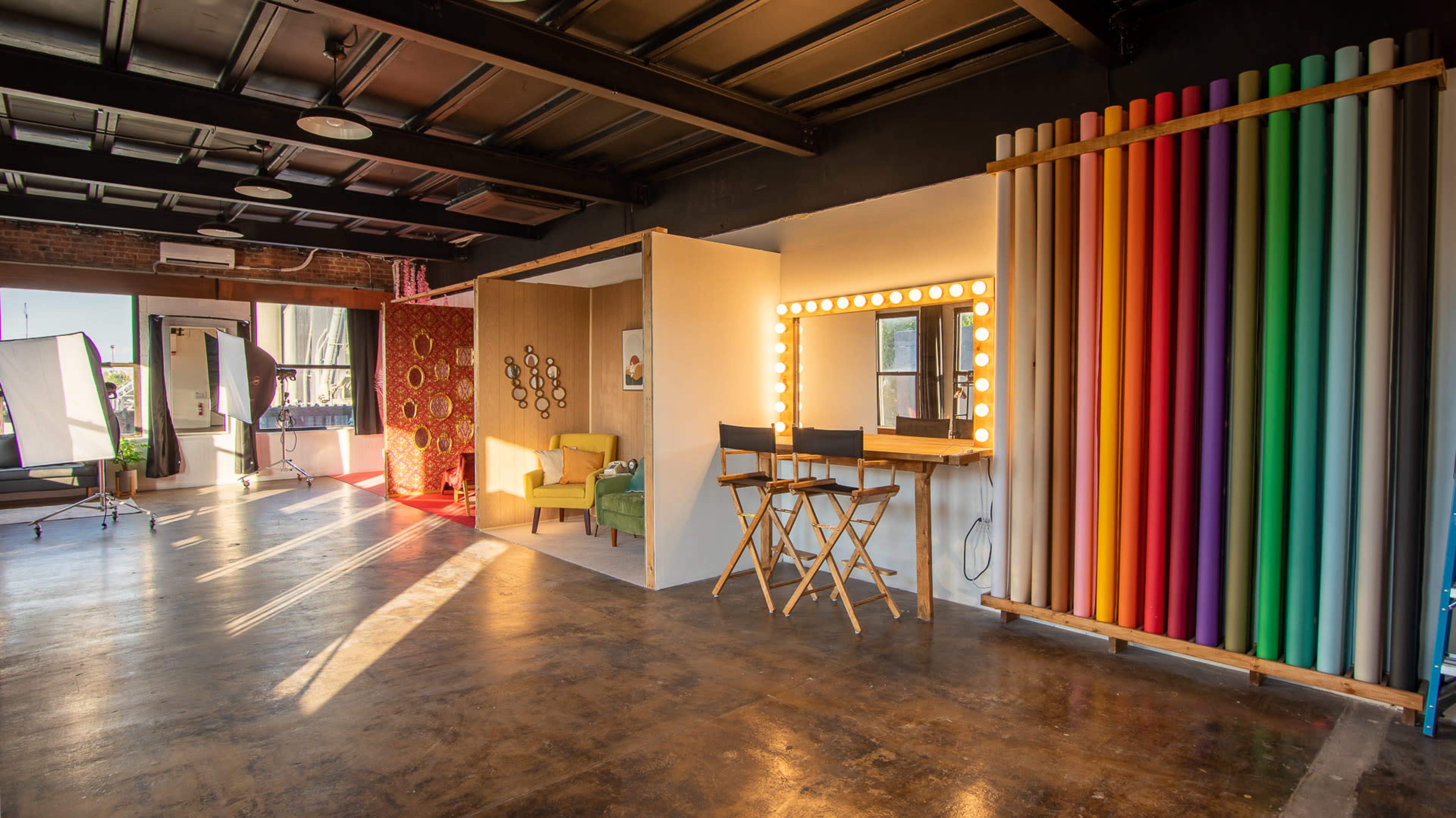 A well-lit studio space featuring various backdrops, a makeup mirror with lights, and director's chairs arranged in an organized layout.