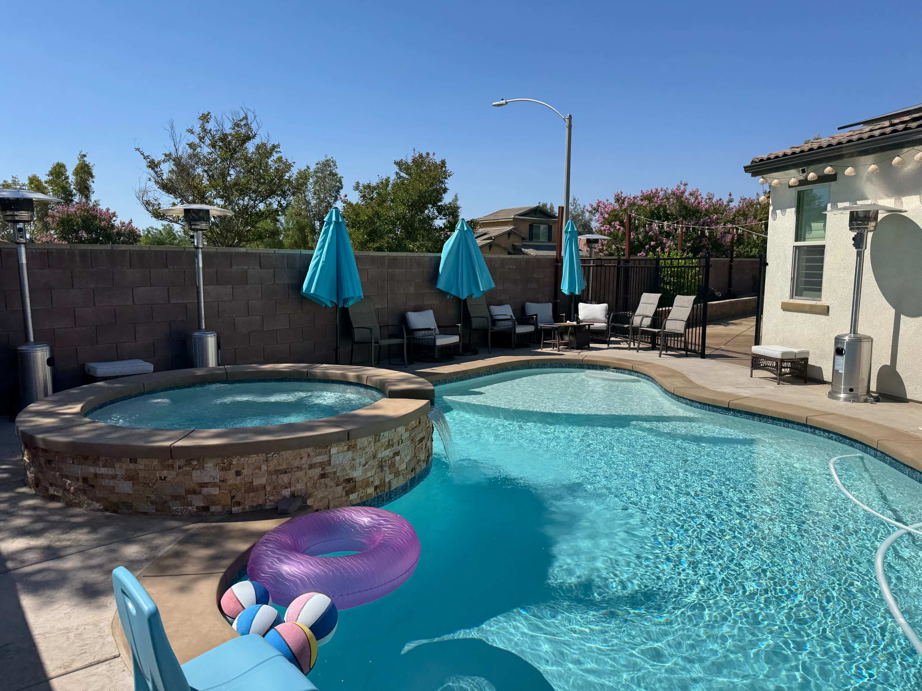 The image shows a backyard pool area featuring a spa, lounge chairs, decorative umbrellas, and a pink inflatable float.