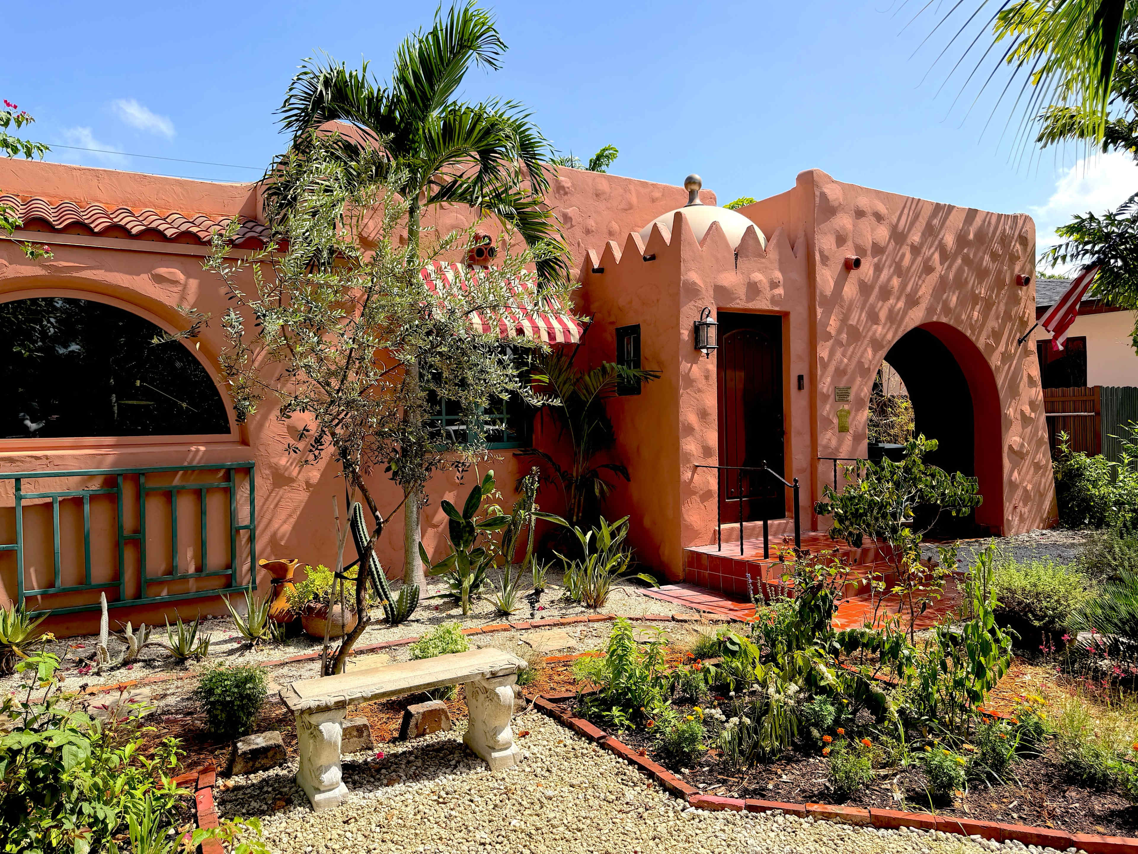 The image shows a colorful, stucco house with arched doorways, a tiled roof, and a garden with various plants and a stone bench in the foreground.