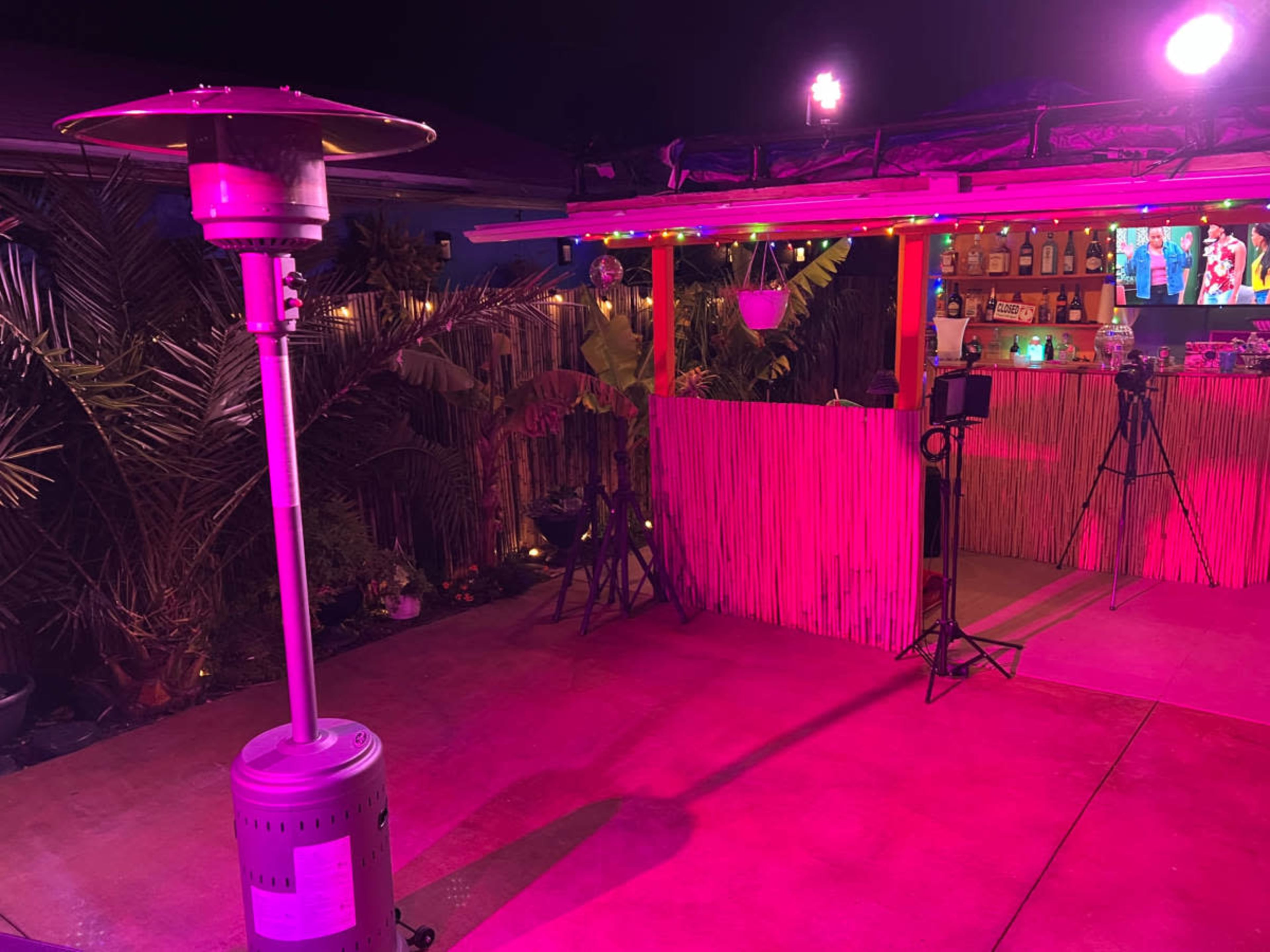 A brightly lit outdoor space with a patio heater, colorful lights, and a bar area surrounded by tropical plants.