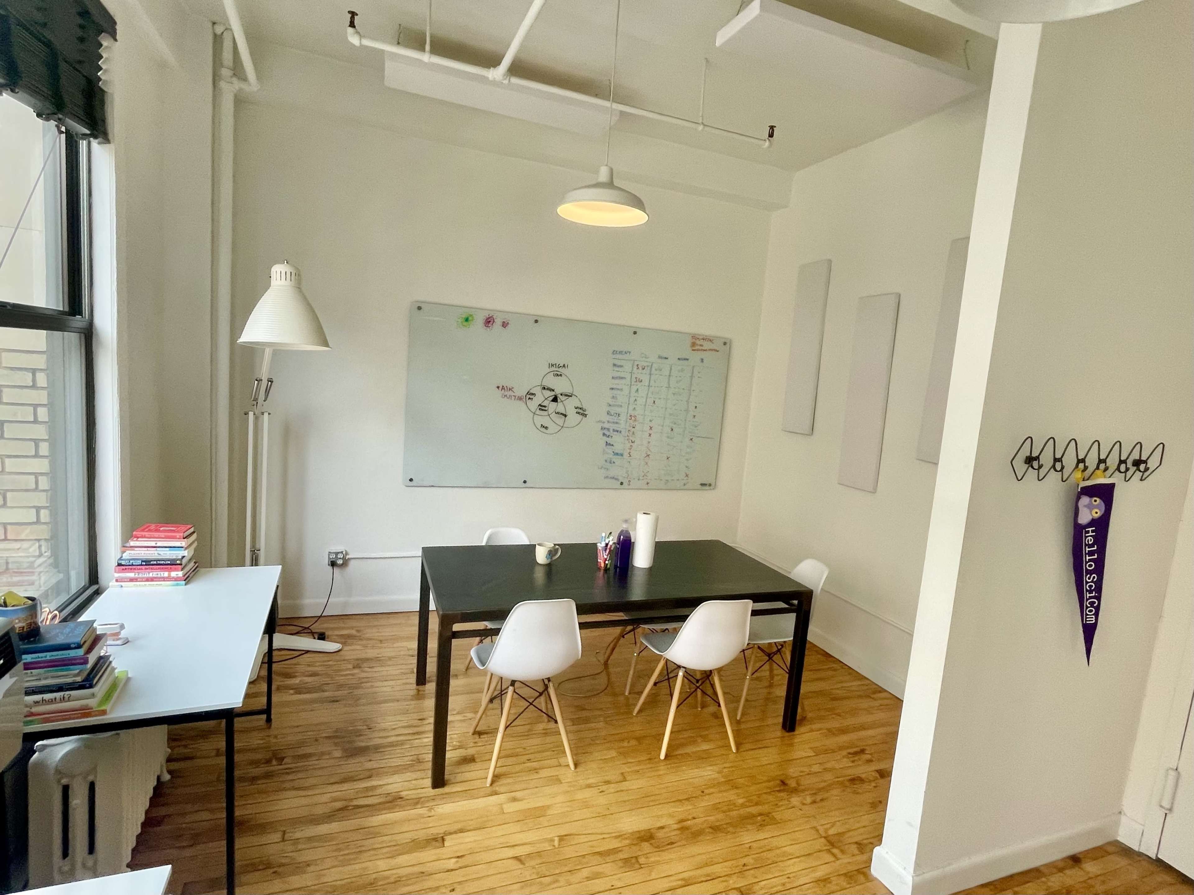 A bright, minimalist office space features a black table surrounded by white chairs, a whiteboard on the wall, and a bookshelf filled with books.
