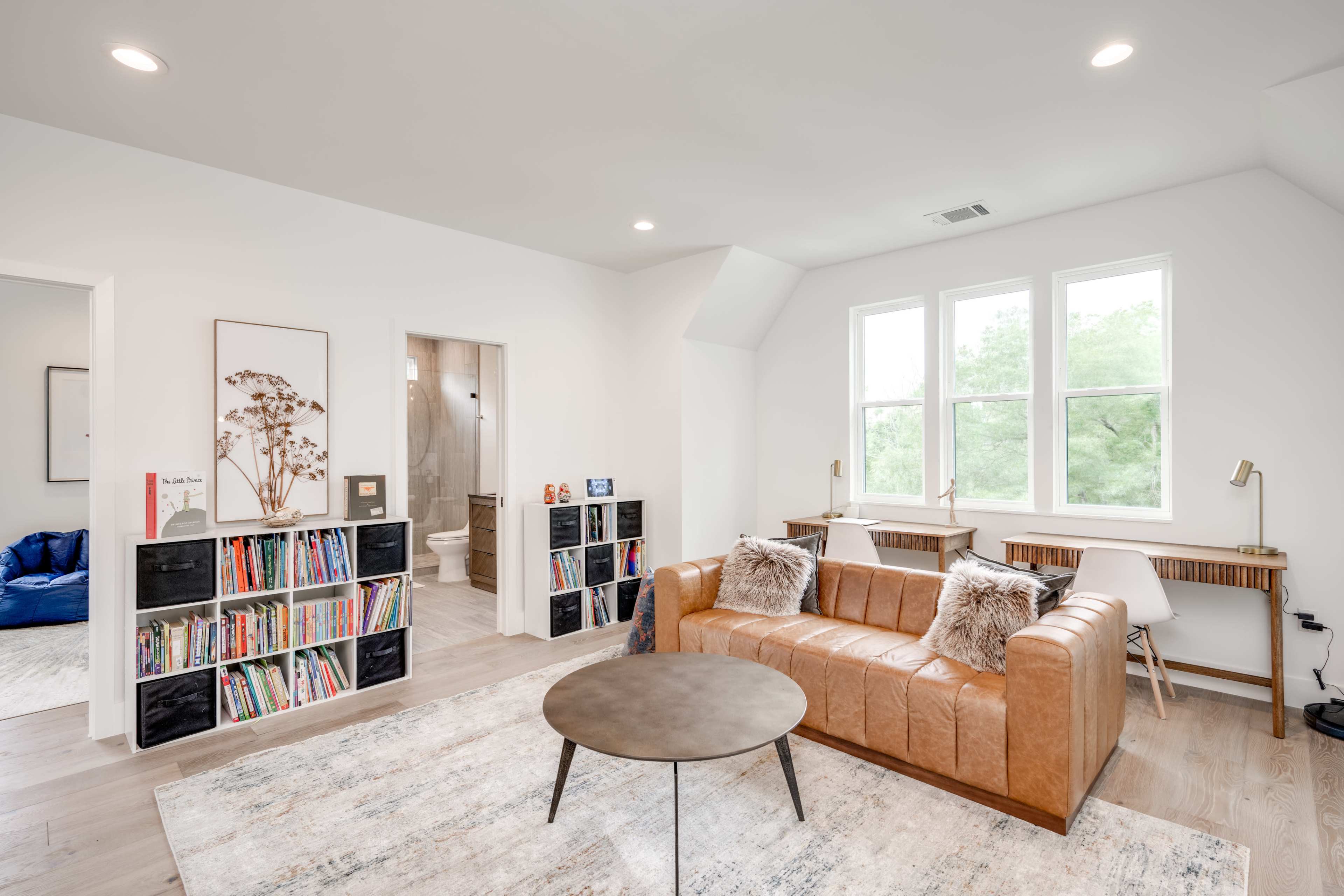 A modern living space features a leather sofa, a bookcase filled with books, and large windows that overlook a green view.