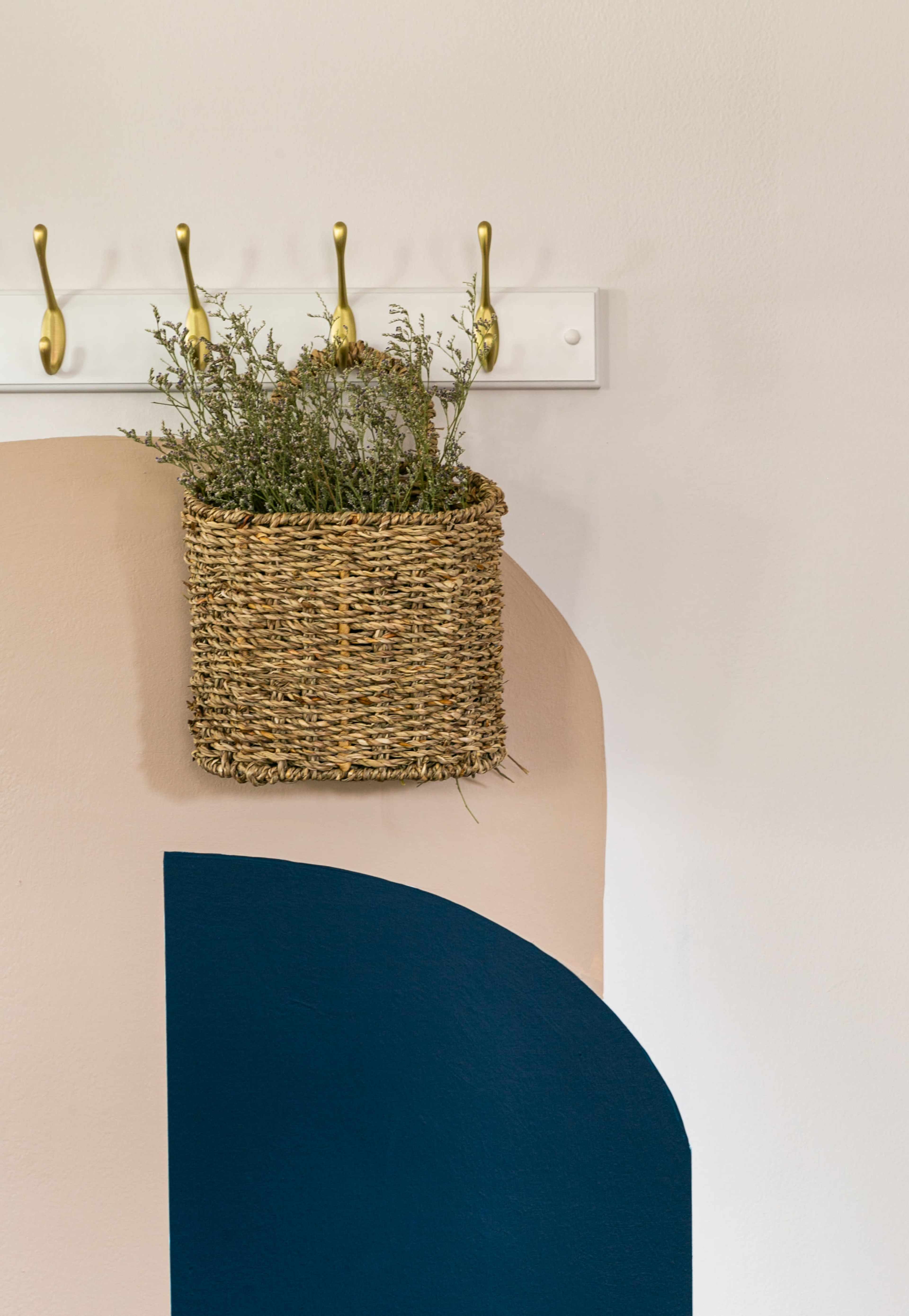 A woven basket filled with greenery is hung on a wall with a hook, next to painted abstract shapes in beige and blue.