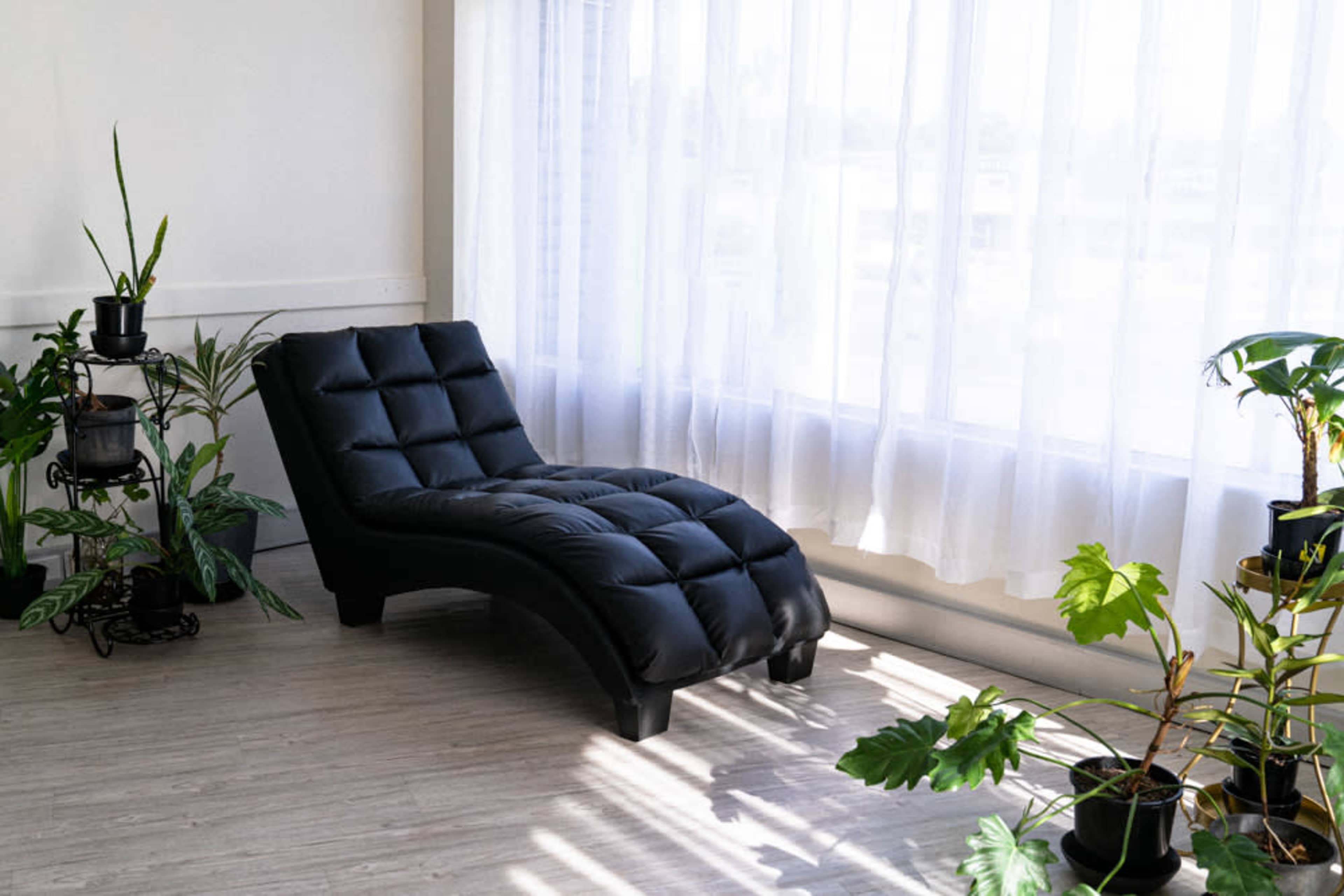 A black chaise lounge is positioned beside sheer white curtains and various potted plants in a light-filled room.