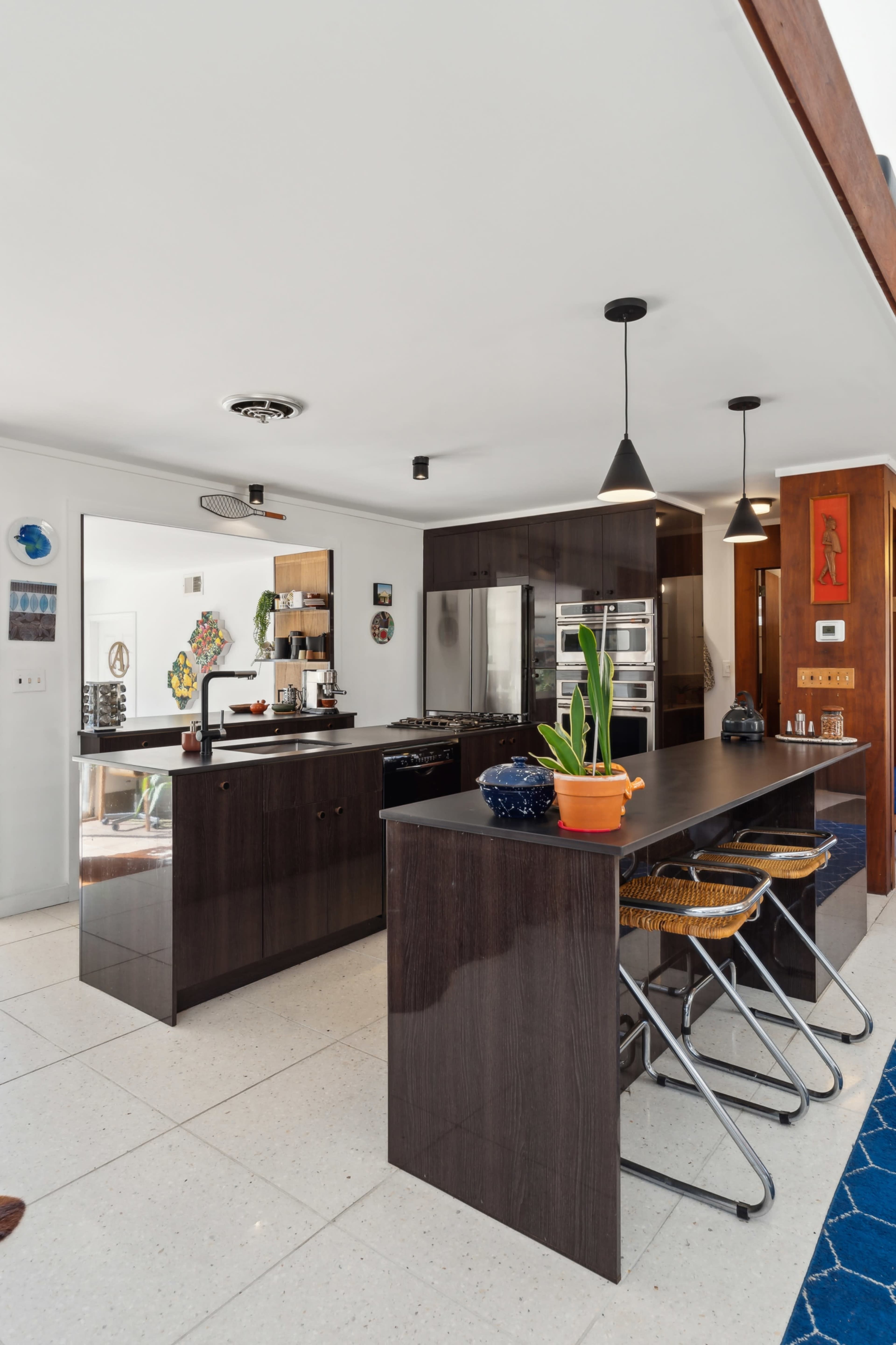 The image shows a modern kitchen with dark cabinetry, a central island, and bar stools, featuring stainless steel appliances and a sleek design.