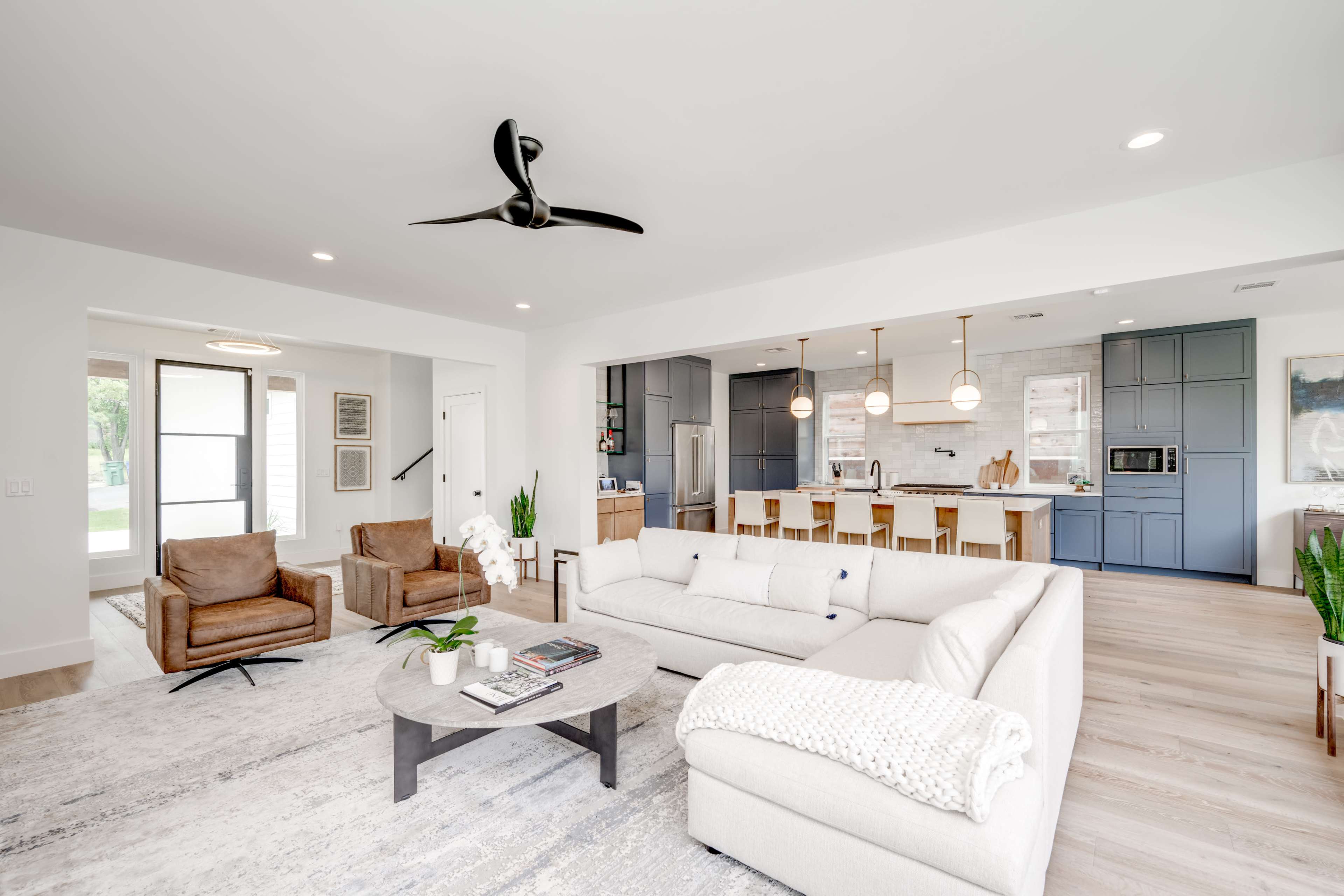 A modern living room features a light-colored sectional sofa, two brown chairs, a round coffee table, and a kitchen area with blue cabinetry and pendant lights.