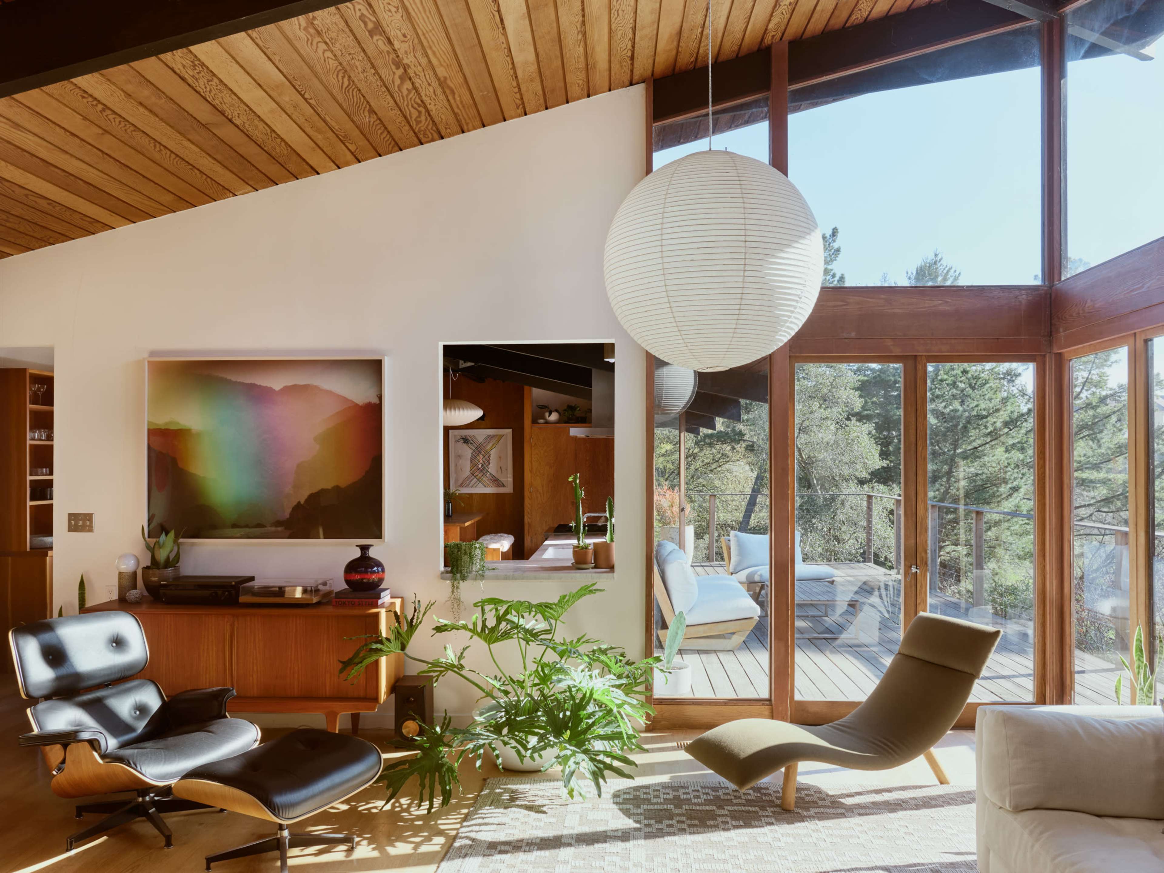 A modern living room features large windows, showcasing a view of trees, with a mix of contemporary furniture and plants.