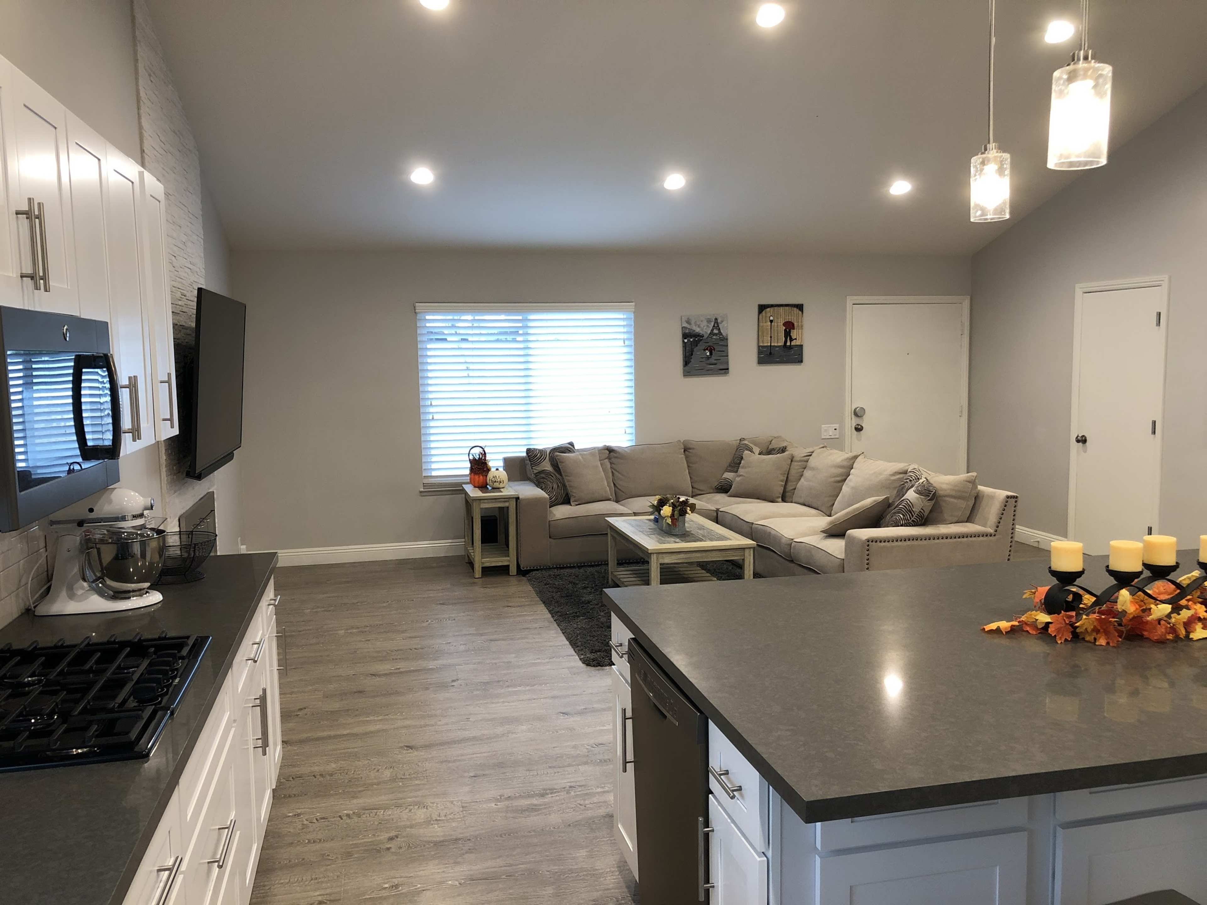 A modern living space features a gray couch, a wooden dining table, pendant lighting, and a kitchen area with white cabinetry and a gas stove.