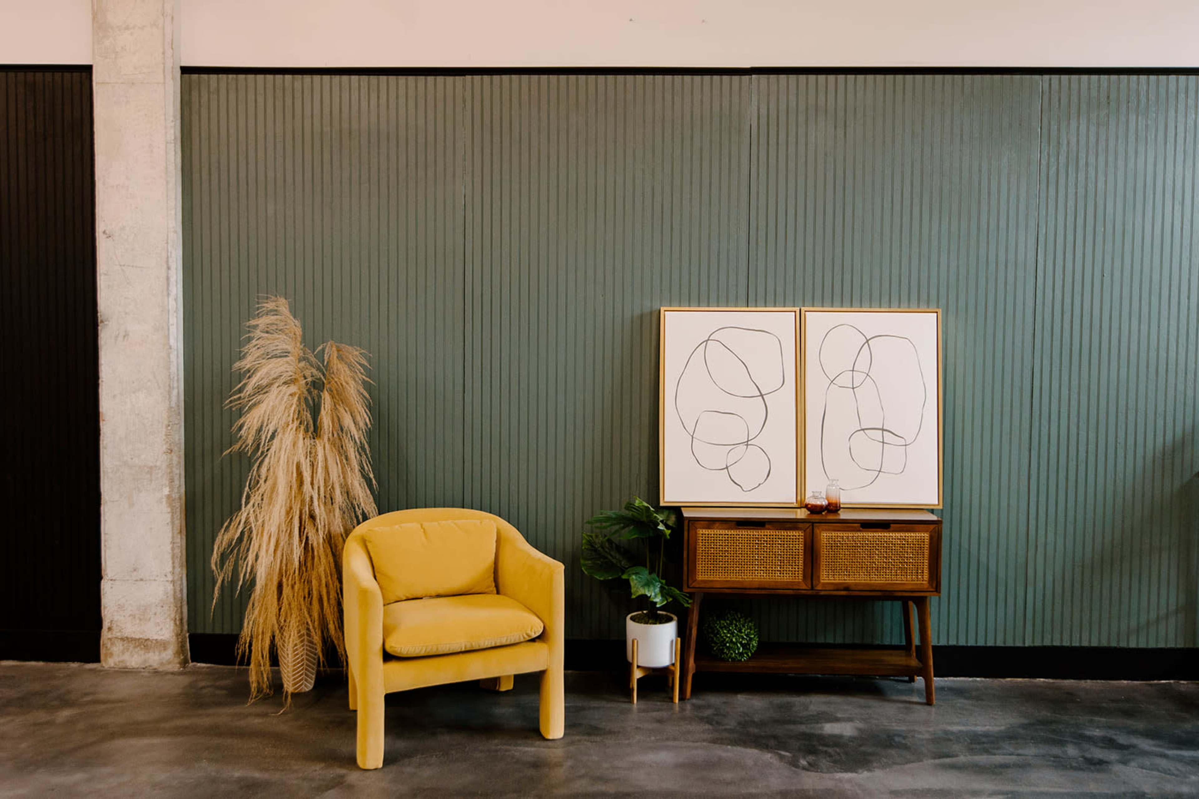 A mustard-colored armchair is positioned next to a wooden console table with two framed abstract line drawings against a green paneled wall, accompanied by a decorative plant and dried grass arrangement.