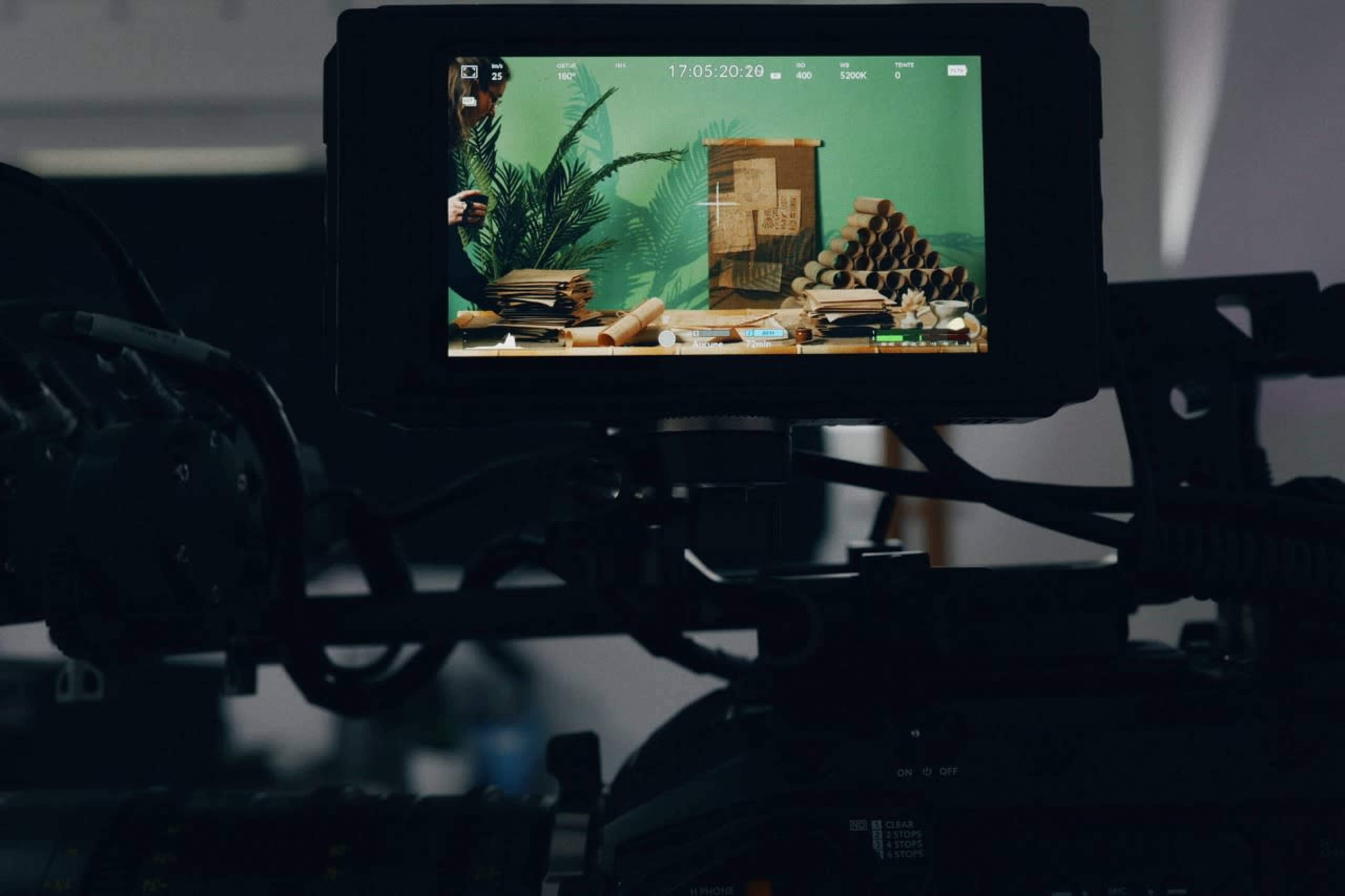 A video camera display shows a set with stacks of books and cylindrical objects against a green background.