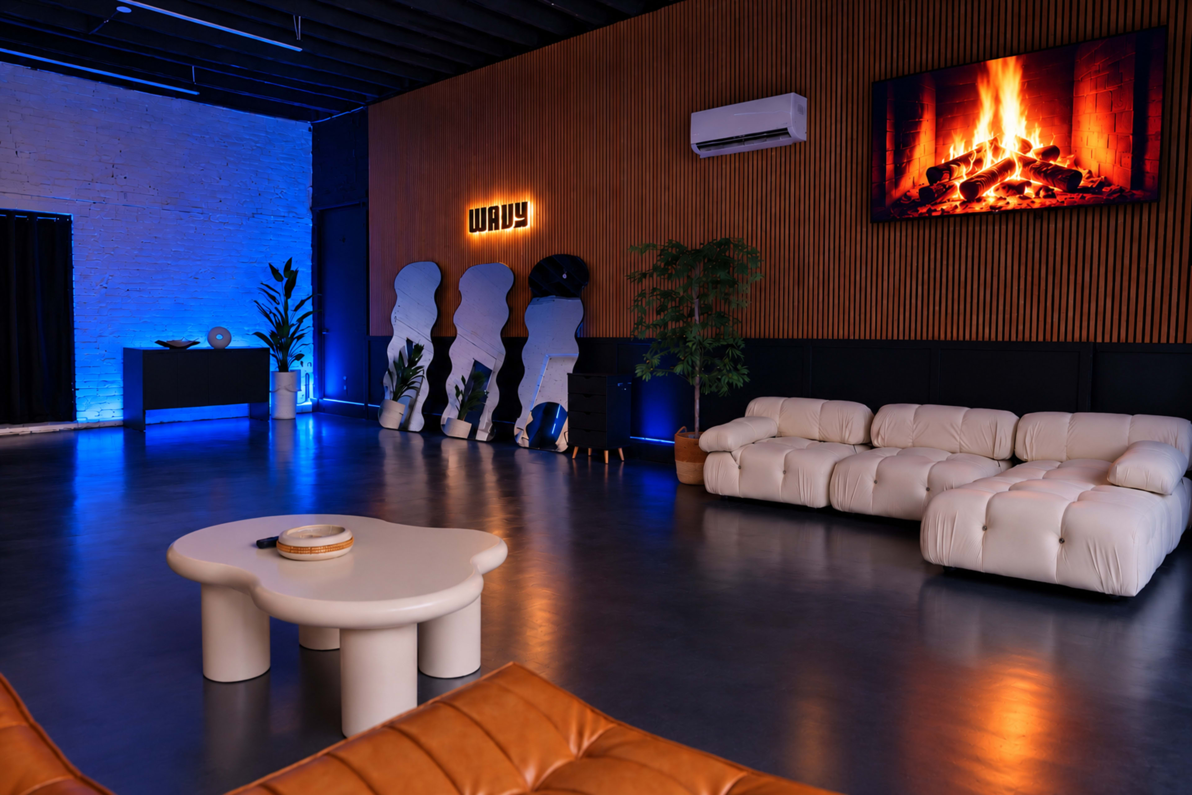 The image shows a modern, minimalist lounge area with a white sofa, a round coffee table, and a wall-mounted television displaying a fireplace, complemented by blue lighting.