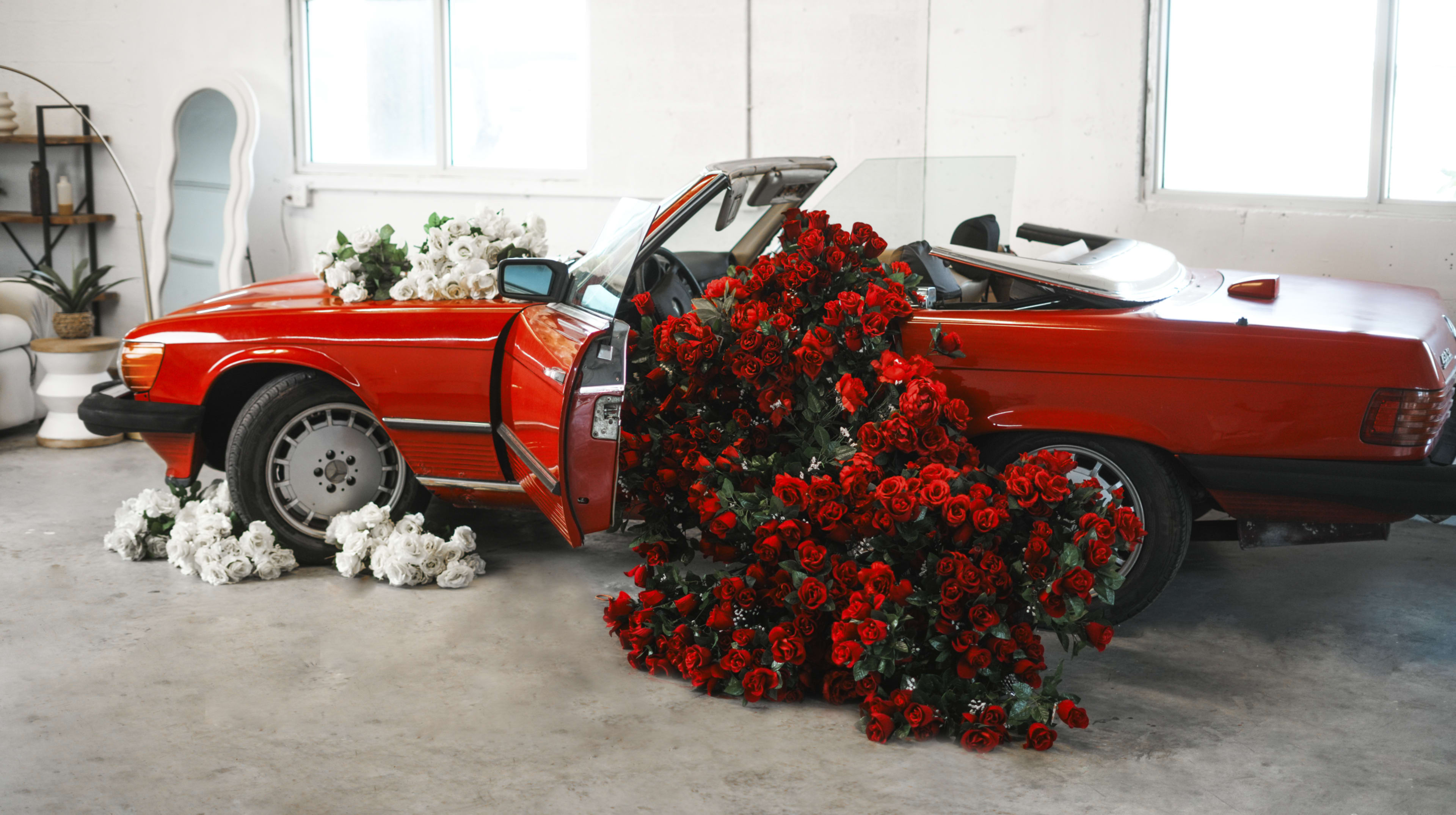 A vintage red convertible car is overflowing with red and white artificial roses, spilling onto the floor in a bright, modern interior space.