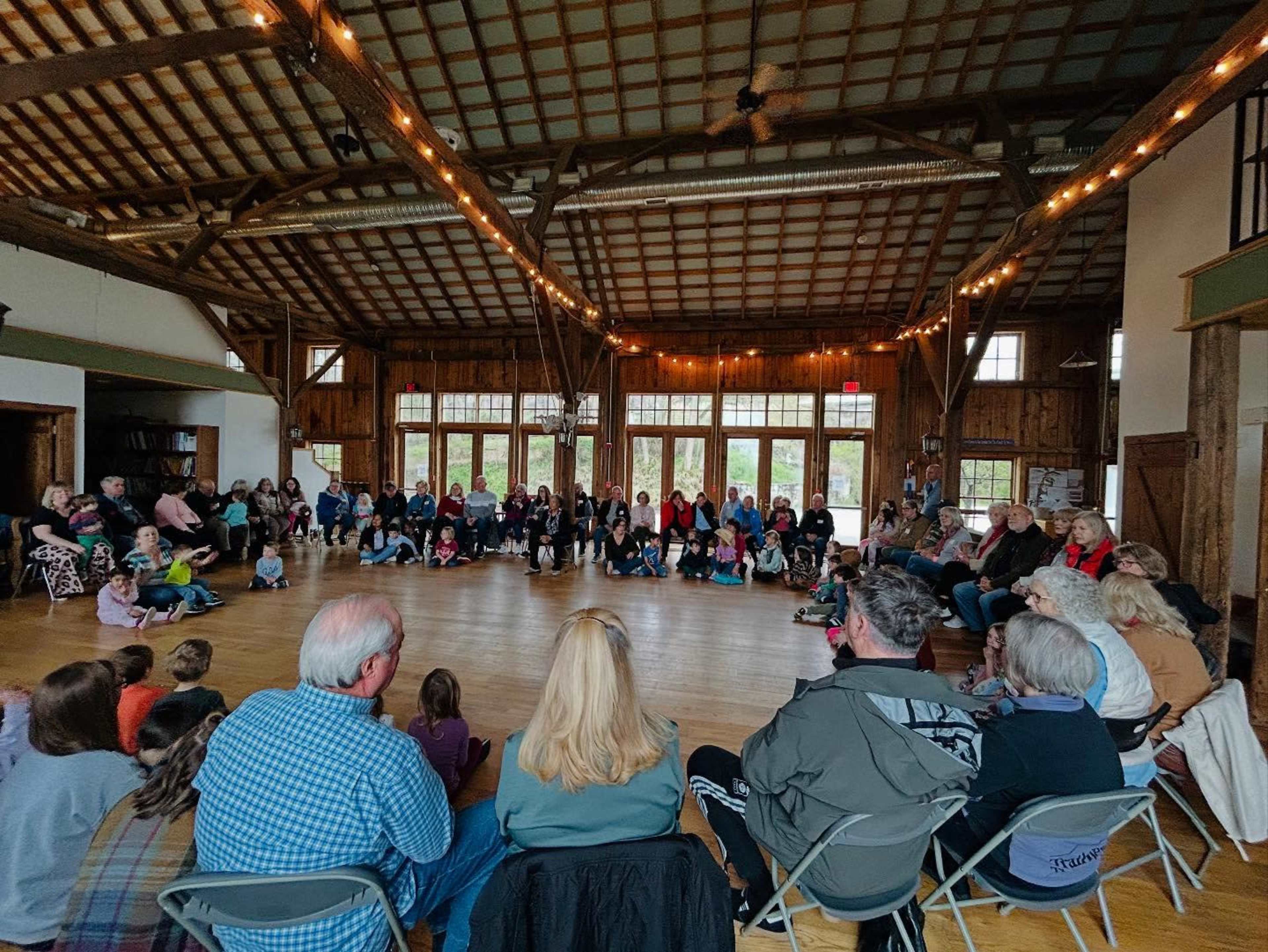 A large group of people sits in a circle on a wooden floor in a spacious hall with high ceilings and large windows.