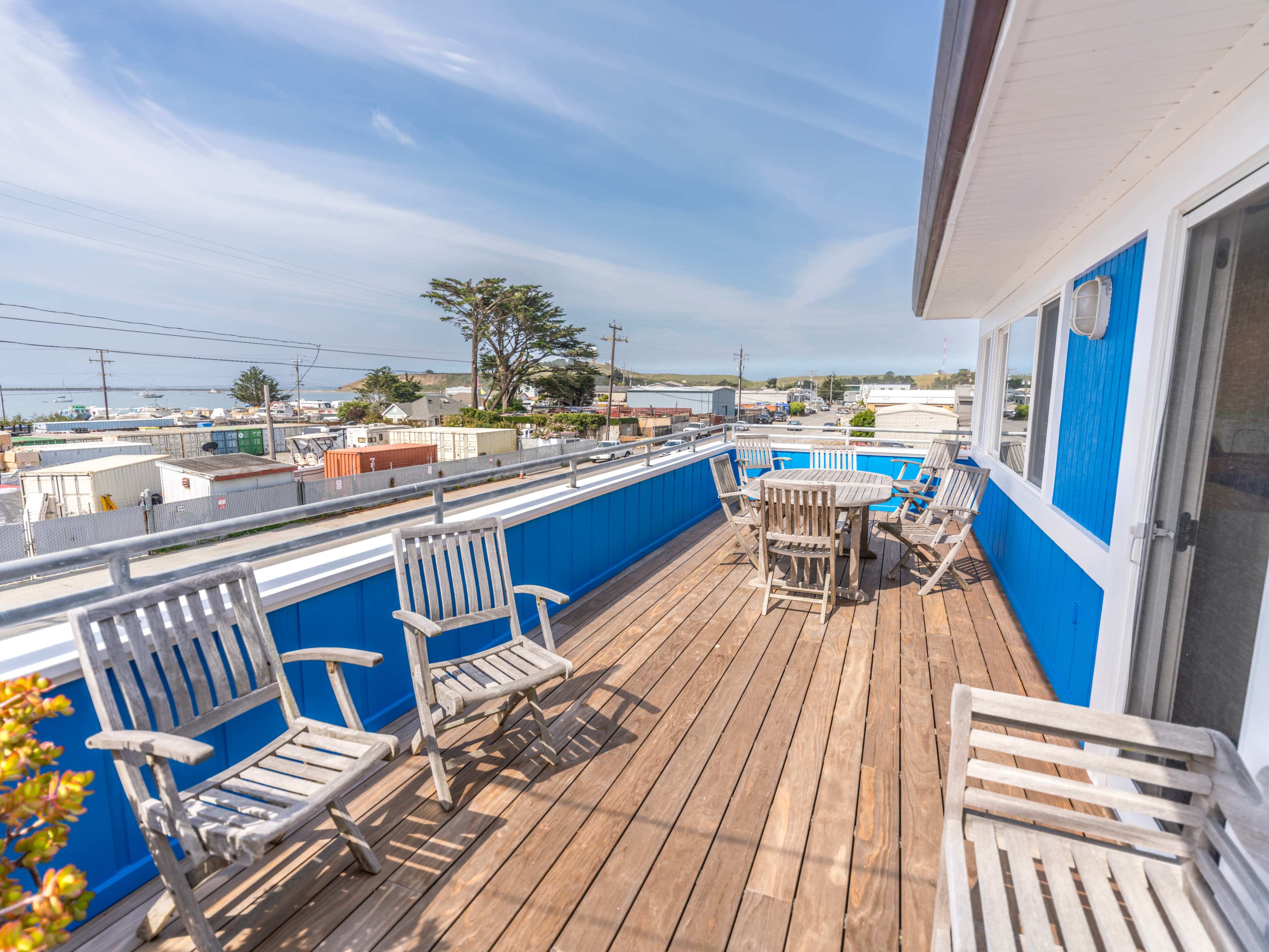 A wooden deck features several chairs and tables with a view of buildings and the ocean in the distance.
