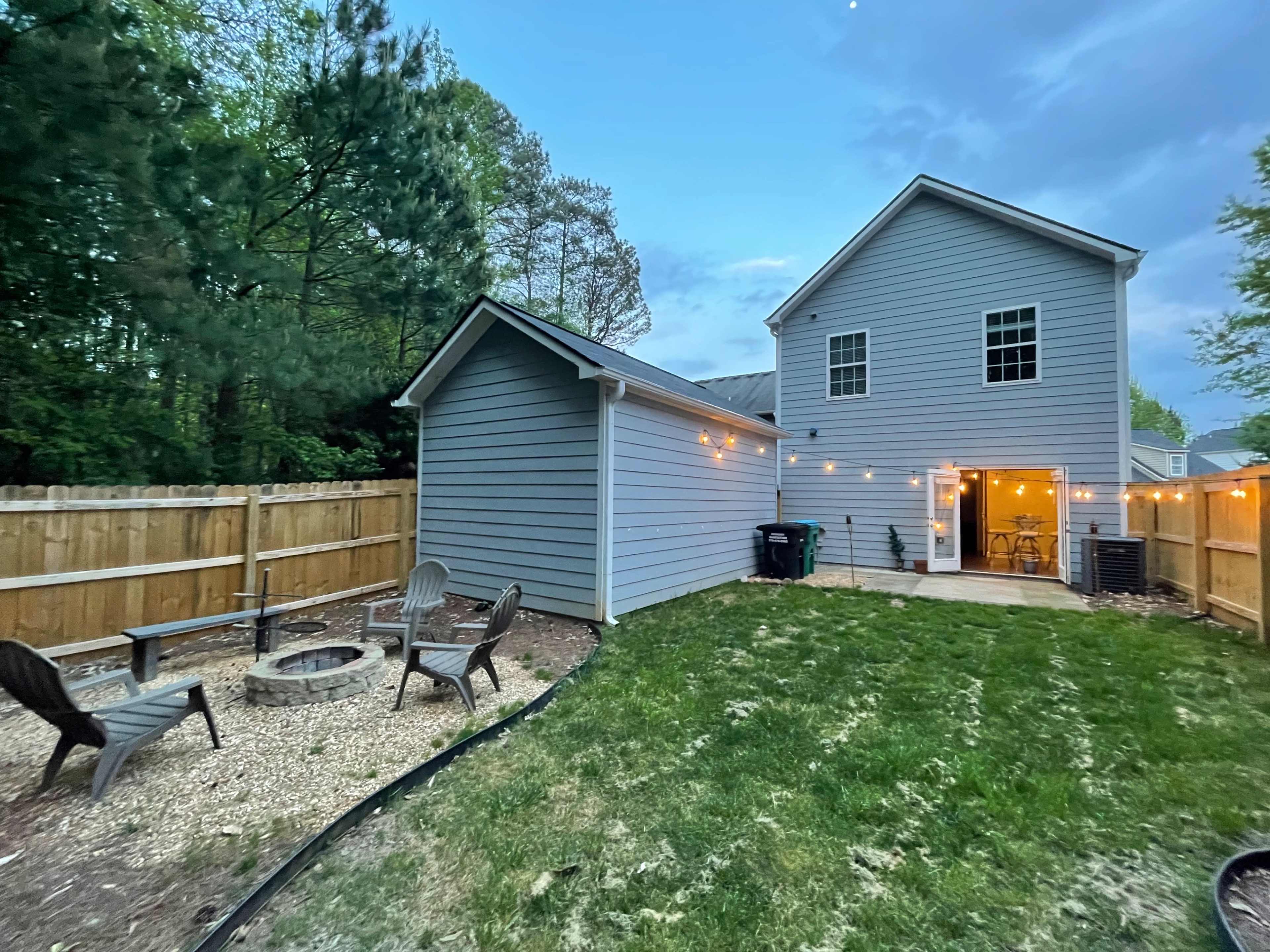 A backyard scene features a fire pit surrounded by chairs, with a two-story gray house and string lights illuminating a patio area.