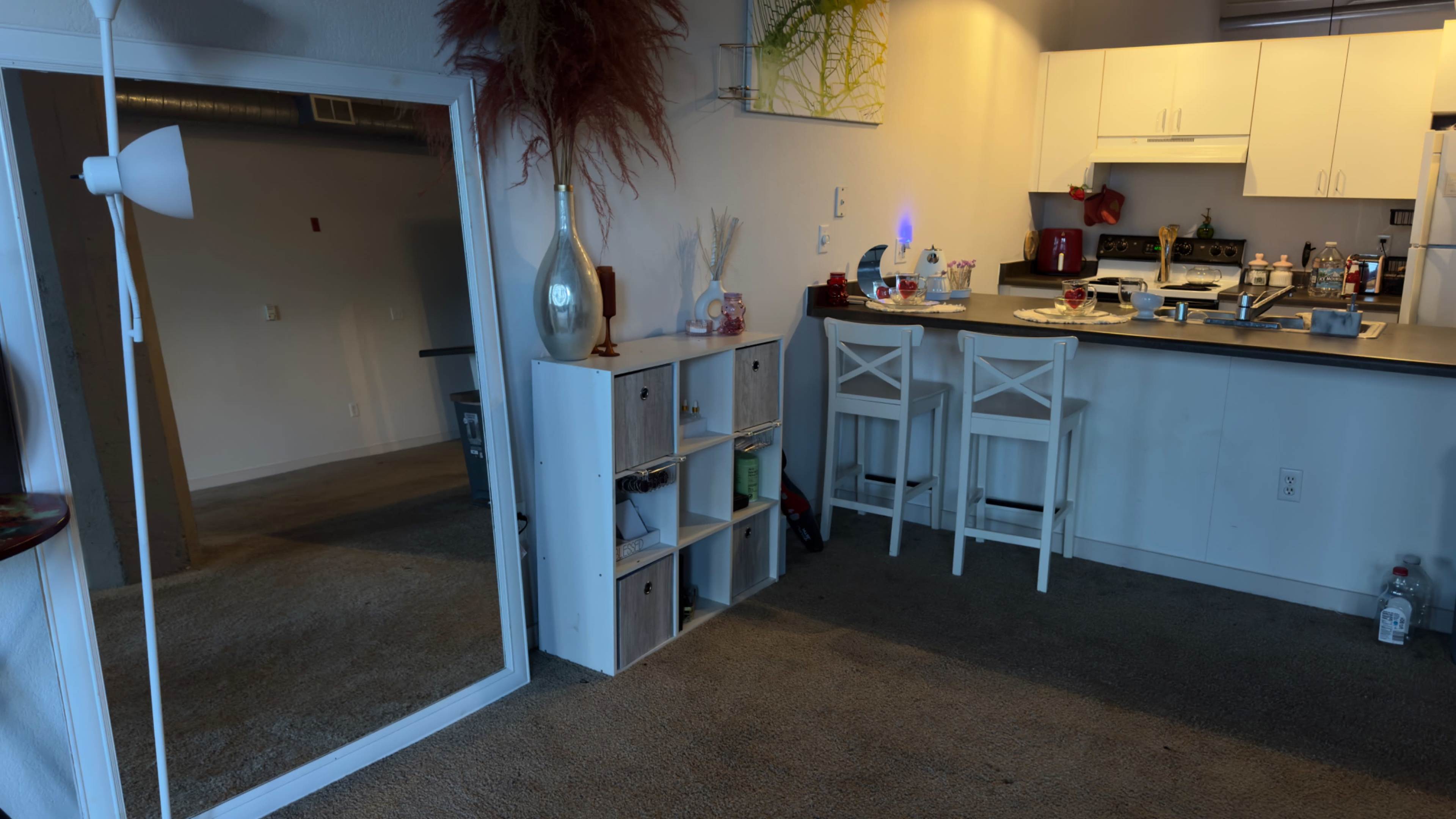 A furnished living area with a large mirror, a white storage unit, and a kitchen counter with stools in a well-lit space.