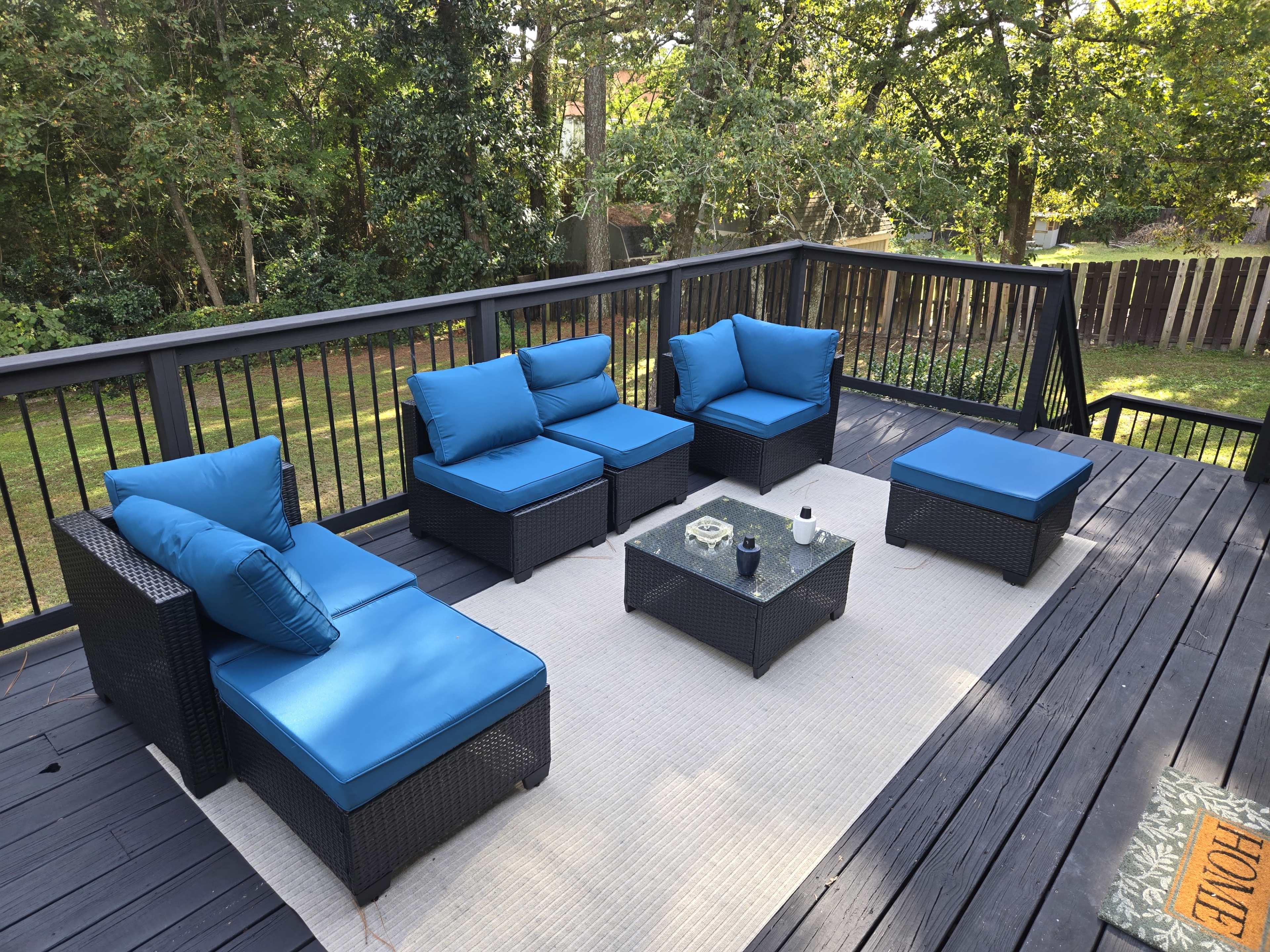 The image shows a deck with a seating area featuring four blue cushioned chairs and two glass-top tables arranged on a wooden surface.