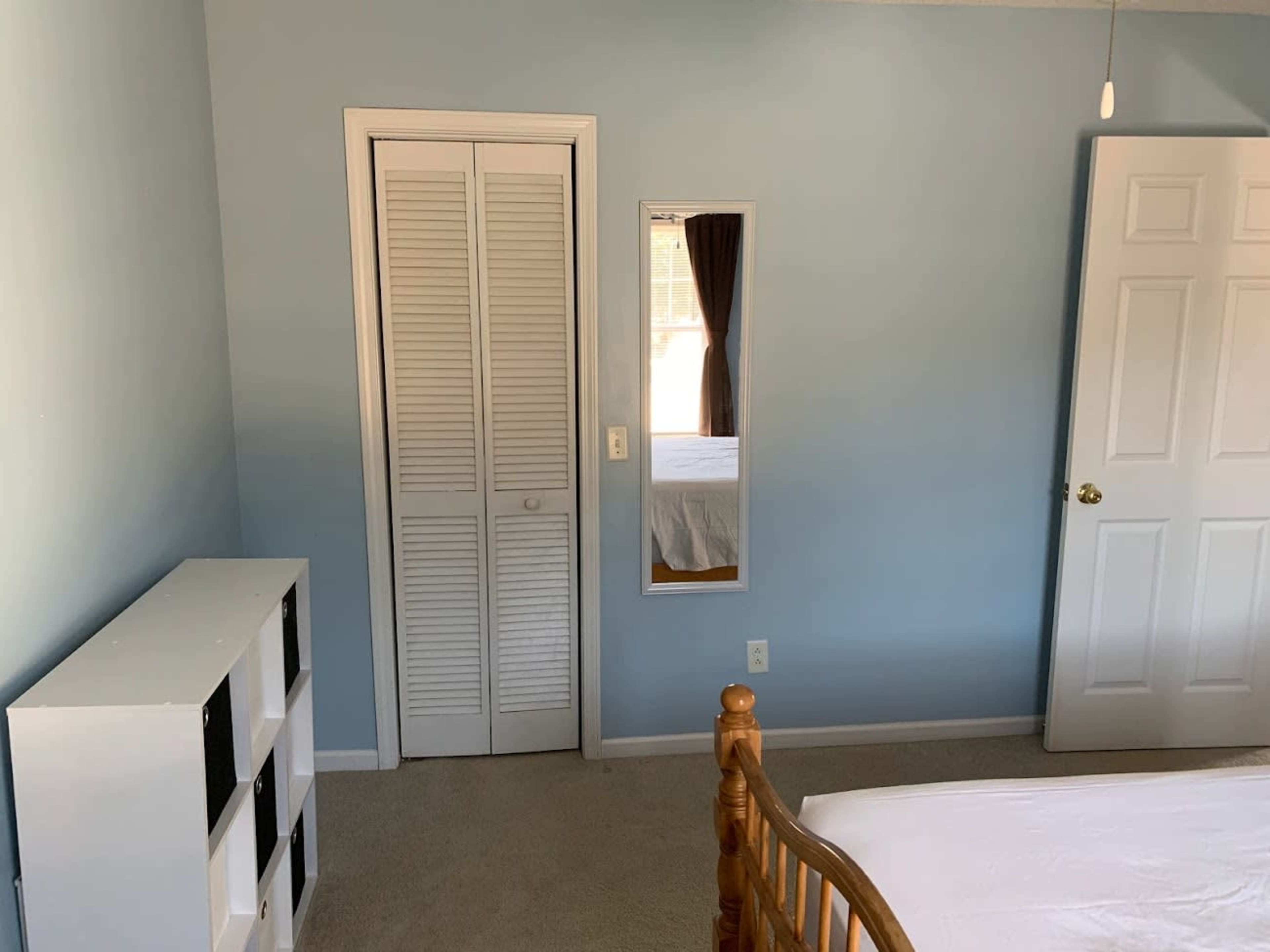 A bedroom features light blue walls, a wooden bed frame, a white bookshelf, and a full-length mirror next to a closet.