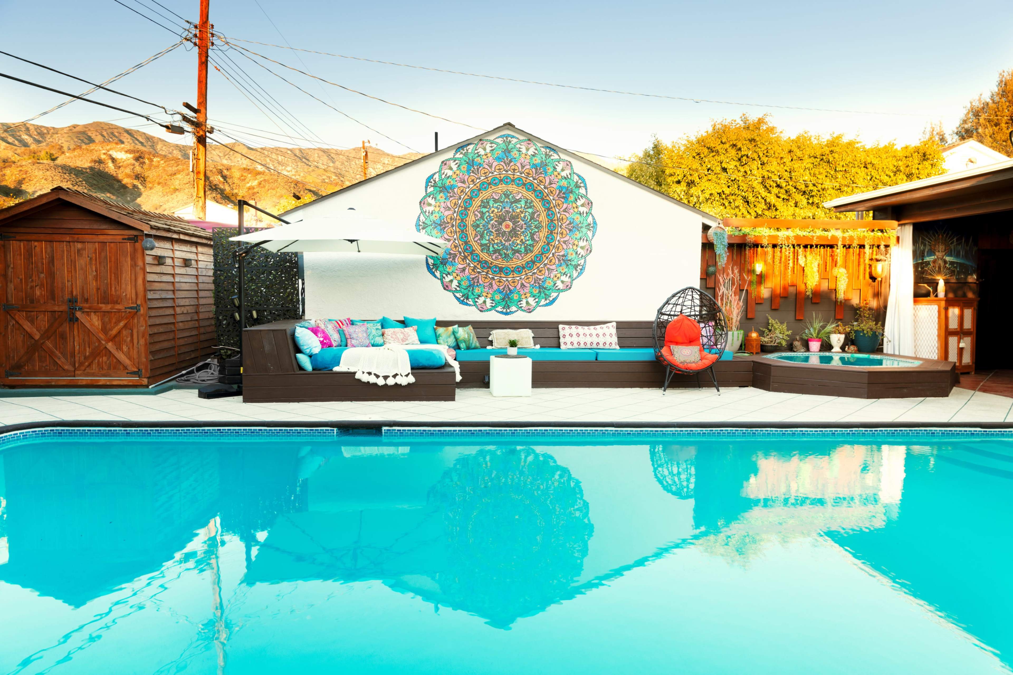 A backyard pool area features a colorful mural on the wall, surrounded by comfortable seating and a red accent chair near the water's edge.