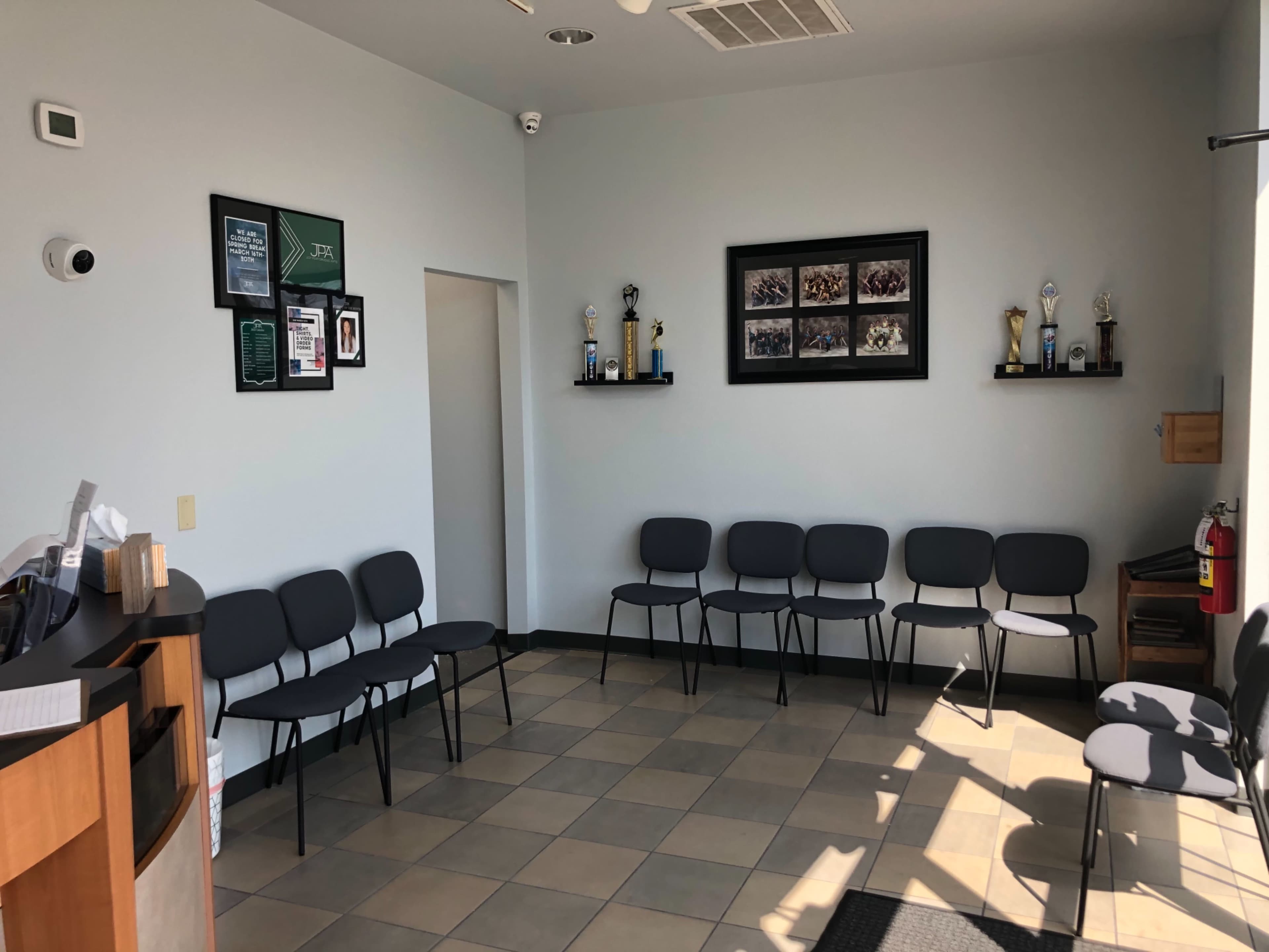 A waiting area with a reception desk, several black chairs arranged in a row, and a wall displaying trophies and framed photographs.