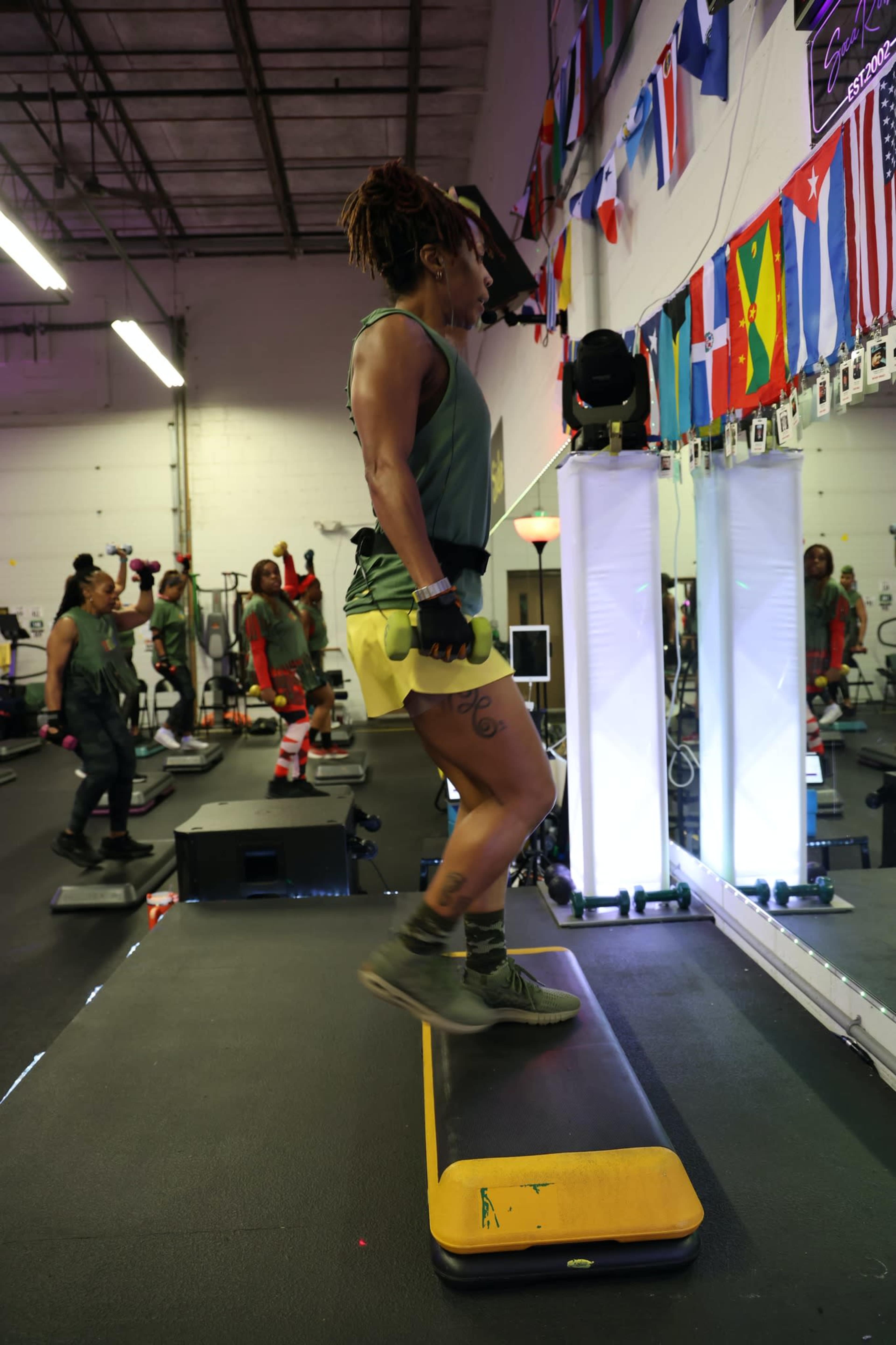 A group fitness class is taking place in a gym, with participants performing exercises on platforms while one individual is jumping and holding weights.