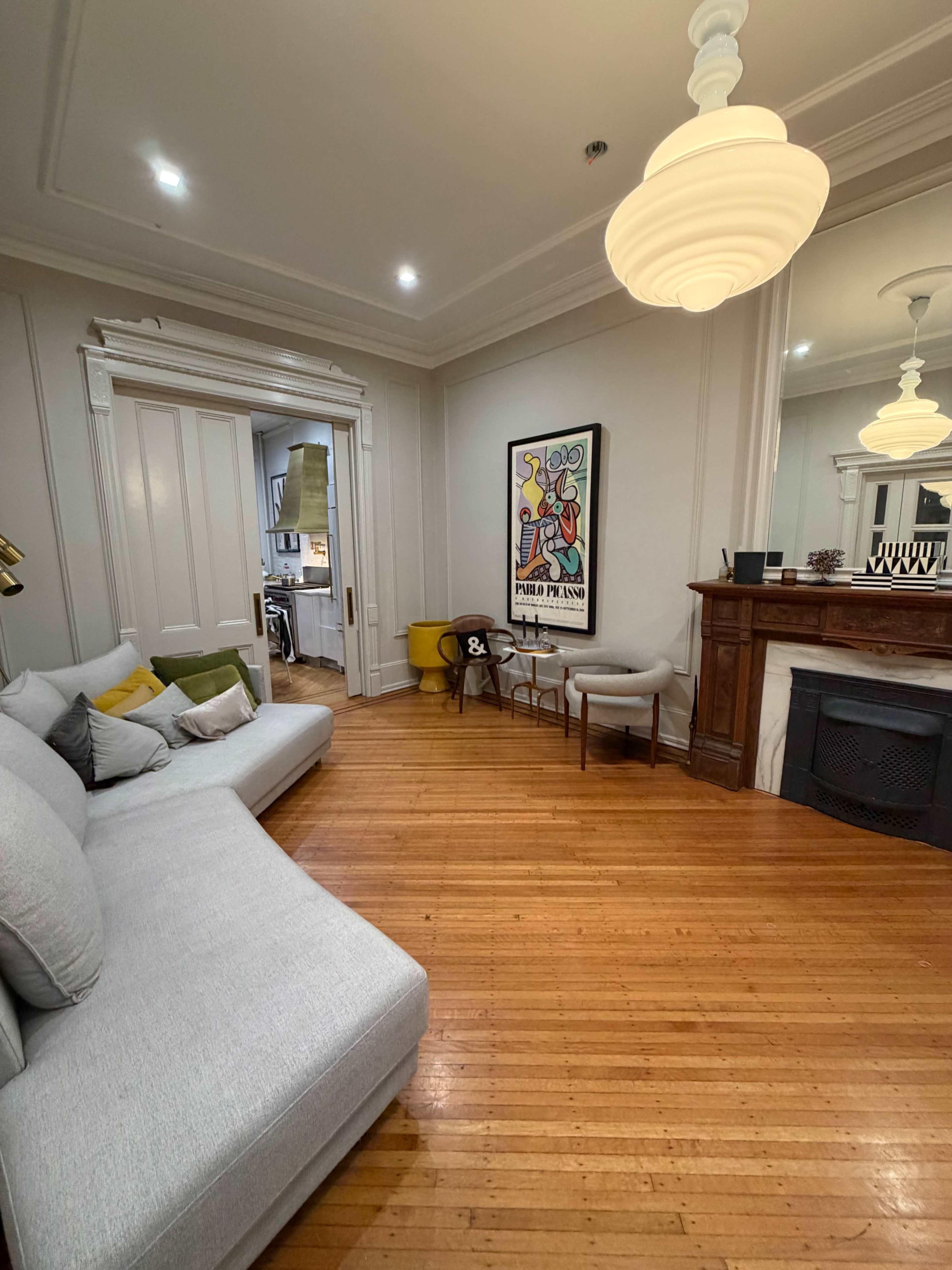 The image shows a living room with a sectional sofa, a small yellow chair, and a framed artwork hanging on the wall, featuring a fireplace and a view into a kitchen area.