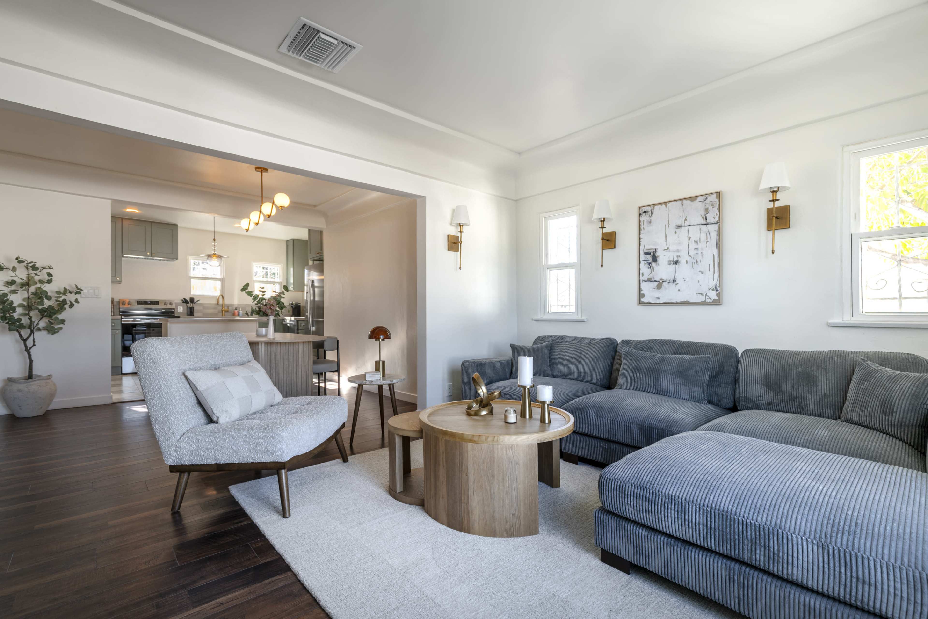 The image shows a modern living room featuring a gray sectional sofa, a round wooden coffee table, and an armchair, with an open view into a kitchen area in the background.