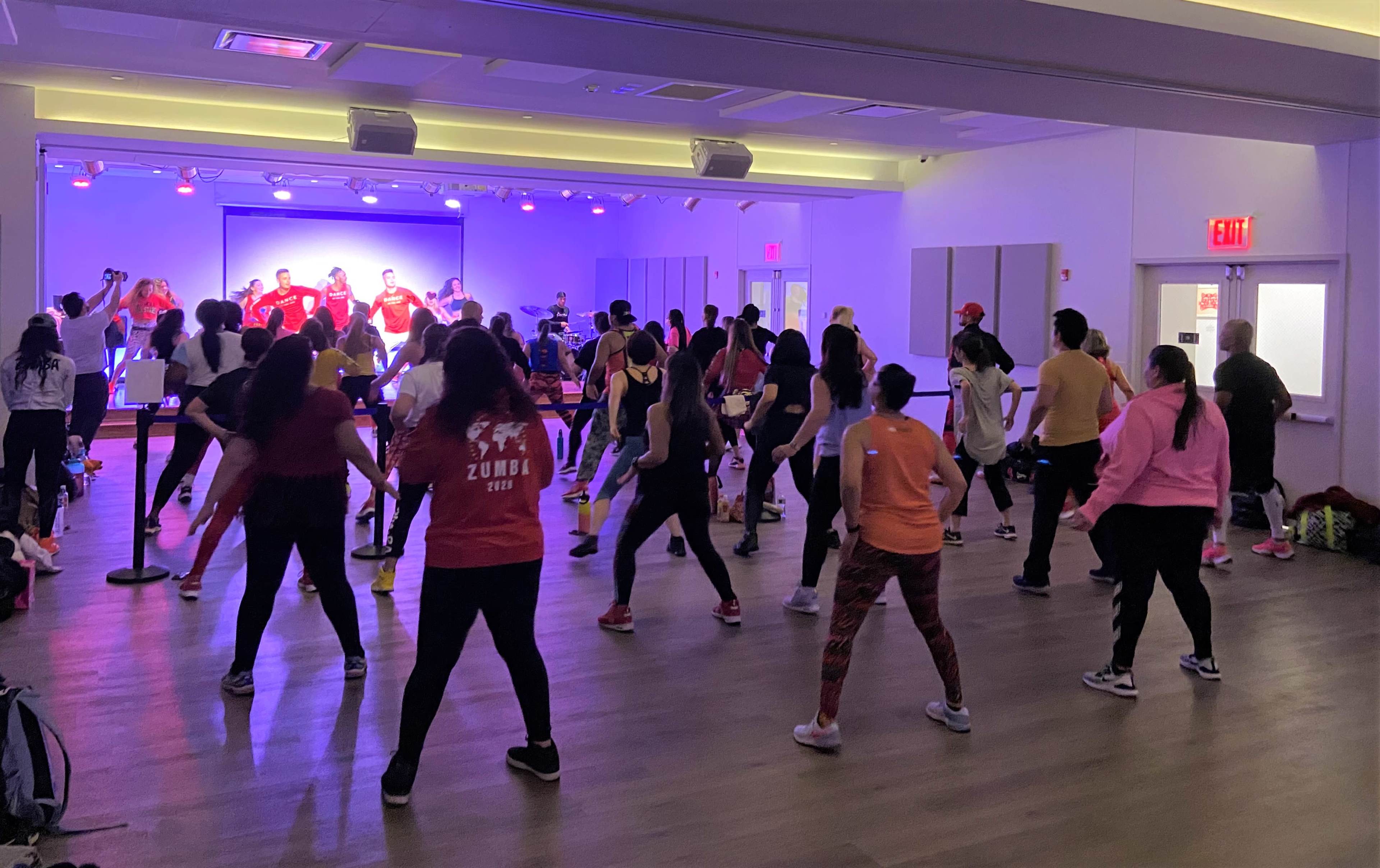 A large group of people are participating in a Zumba class in a brightly lit studio with a stage and colorful lighting.