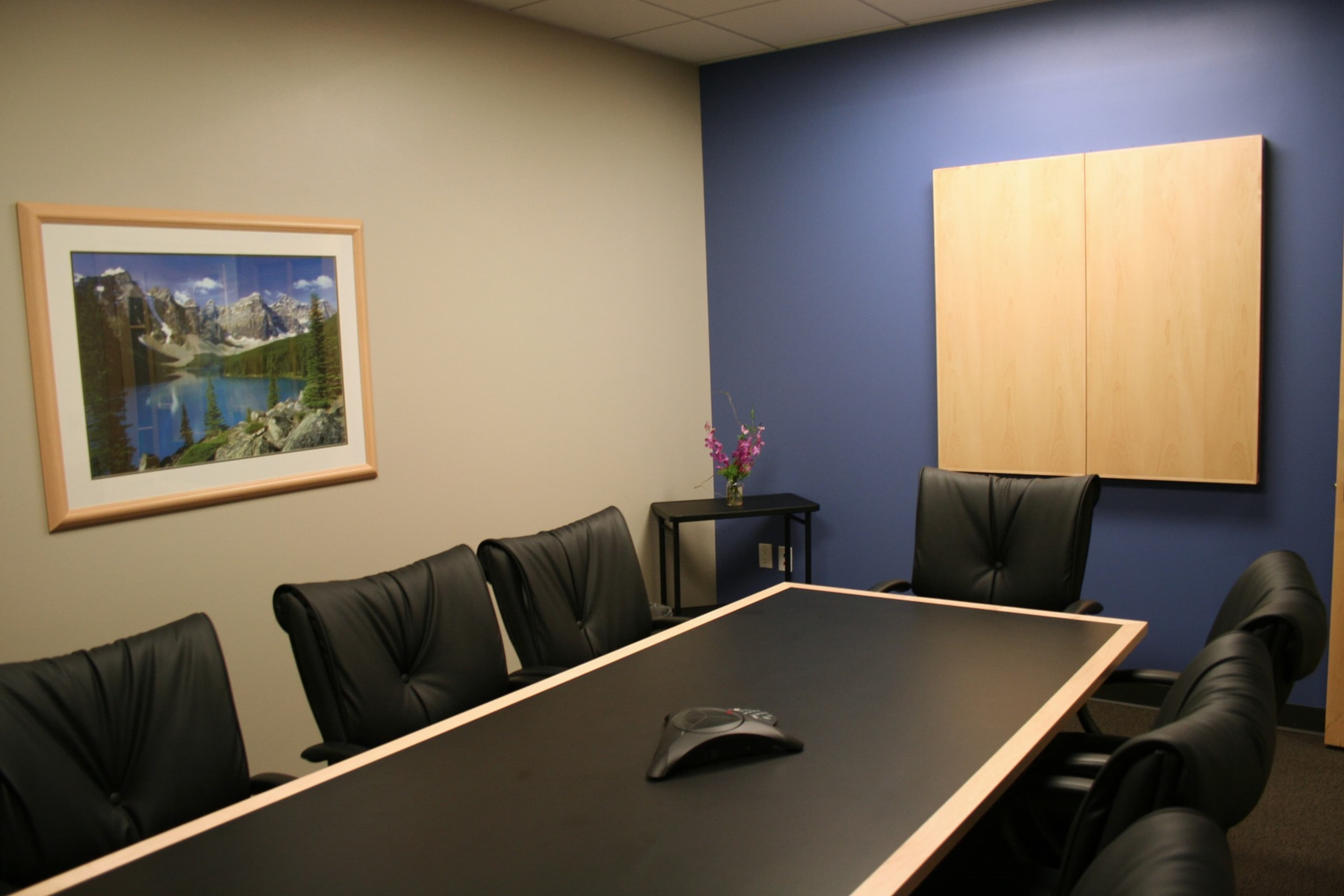 A modern conference room features a long black table surrounded by black leather chairs, with a landscape painting on one wall and a wooden cabinet on another.