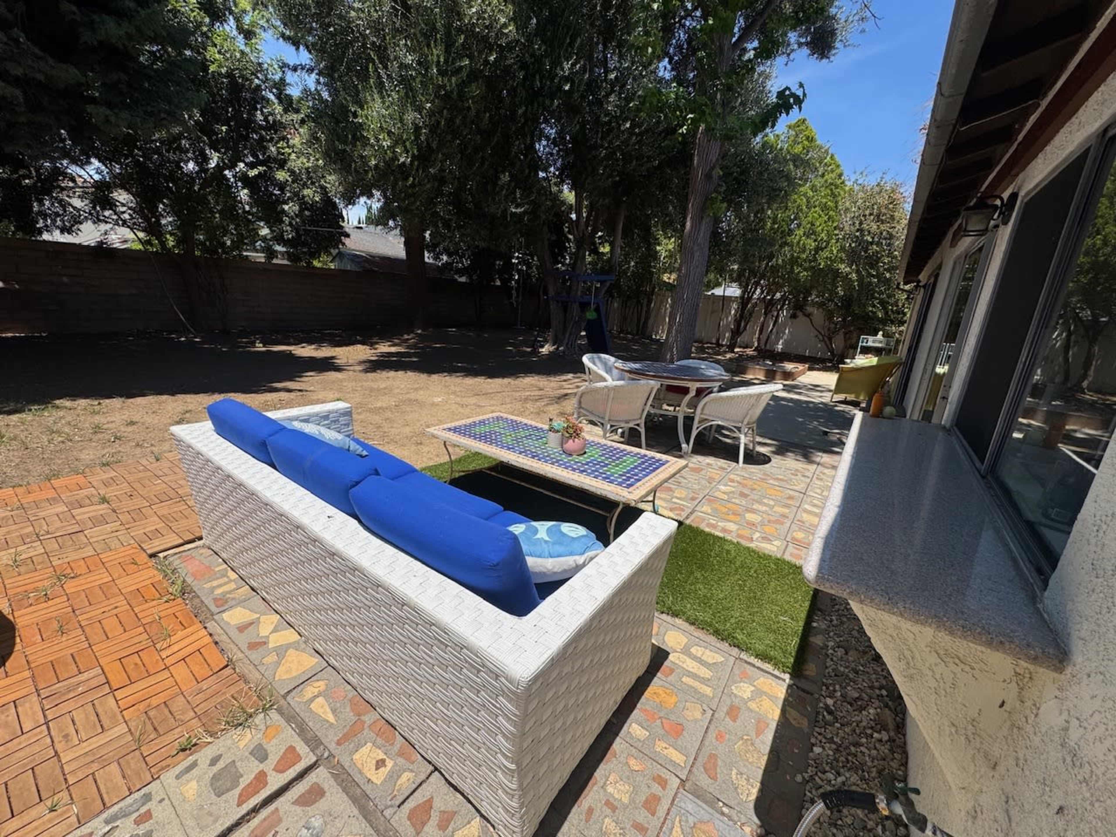 The image shows a backyard with a patio area featuring a white wicker sofa with blue cushions, a small table on a patterned stone surface, and a dirt lawn surrounded by trees.